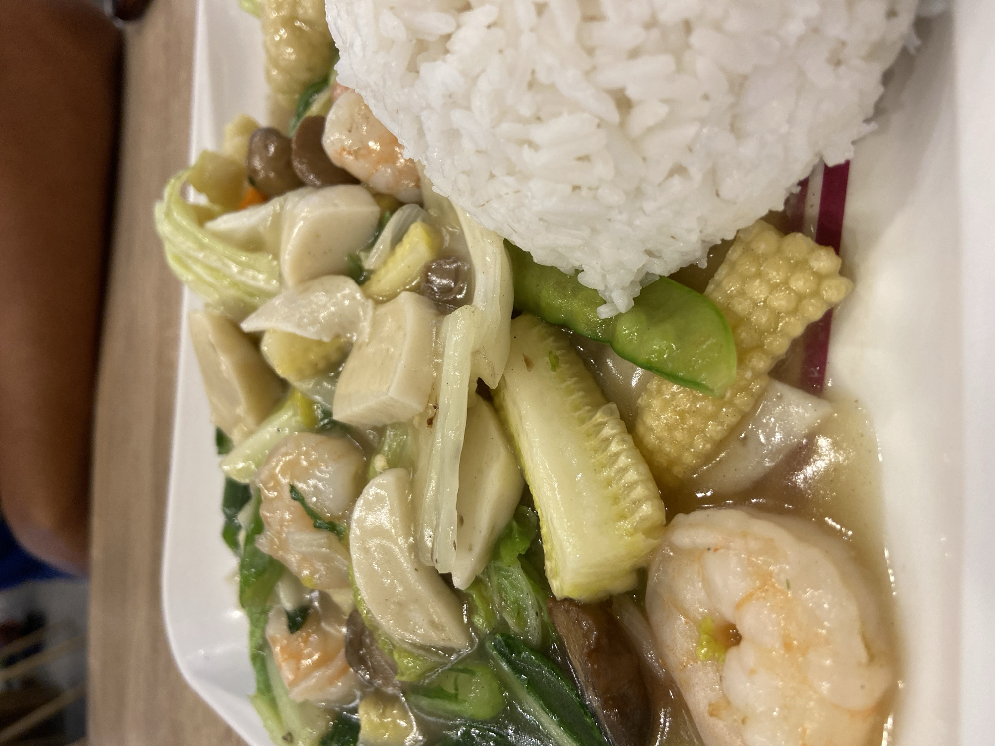 A close-up, eye-level shot shows a dish of stir-fried seafood and vegetables with a side of white rice, served on a white plate. The dish features several large, pink shrimp, chopped bok choy, slices of napa cabbage, baby corn, mushrooms, and what appear to be pieces of fish cake or tofu, all coated in a light, glossy sauce. The rice is piled high on the right side of the plate, with a portion of the stir-fry visible in front of it. The background is blurred, but it suggests an indoor setting with a wooden table and a person's arm and shoulder visible. The lighting is bright, highlighting the textures and colors of the food. This dish is likely from a restaurant in Sidoarjo, Indonesia, given the common culinary style.