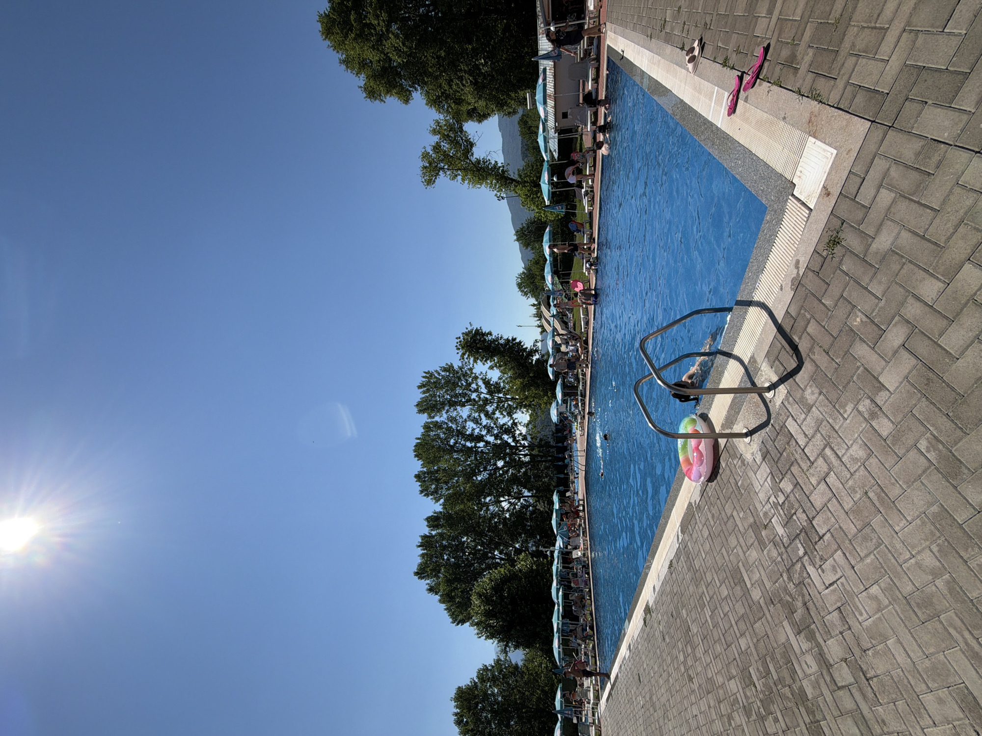 This image depicts a bright, sunny day at an outdoor swimming pool complex, likely located in Simitli, Bulgaria. The foreground is dominated by a paved area made of grey interlocking bricks, which leads to the edge of a large, rectangular swimming pool filled with clear blue water. A set of stainless steel pool stairs with handrails is partially submerged in the water on the right side of the frame. Near the stairs, a colorful inflatable ring floats on the surface of the water.
On the opposite side of the pool, numerous lounge chairs with open umbrellas are arranged, indicating an area for sunbathing and relaxation. Several people are visible around the pool, some are in the water swimming or wading, while others are seated or standing on the deck. The pool area is bordered by lush green trees and foliage, with mountains visible in the background under a clear blue sky. The sun is high in the sky, casting bright sunlight and creating reflections on the water's surface. Scattered on the paved area near the pool's edge are a few pairs of flip-flops. The scene suggests a recreational environment, possibly a public pool or a resort.