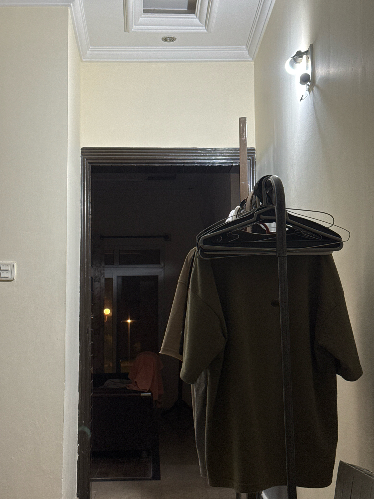 This image captures an indoor hallway scene, likely within a residential or domestic setting in Kohistan Enclave, Pakistan. A doorway, framed by dark wood, leads into a dimly lit interior space. To the right of the doorway, a coat rack holds several black hangers, one of which is draped with a dark green oversized garment, possibly a t-shirt or sweater. The wall to the right of the coat rack features a light fixture with two exposed bulbs, casting a warm glow. The wall to the left is painted a light cream color and has a white light switch panel. The ceiling displays detailed molding and a recessed square light fixture. The overall ambiance is one of a functional, perhaps slightly cluttered, domestic interior.