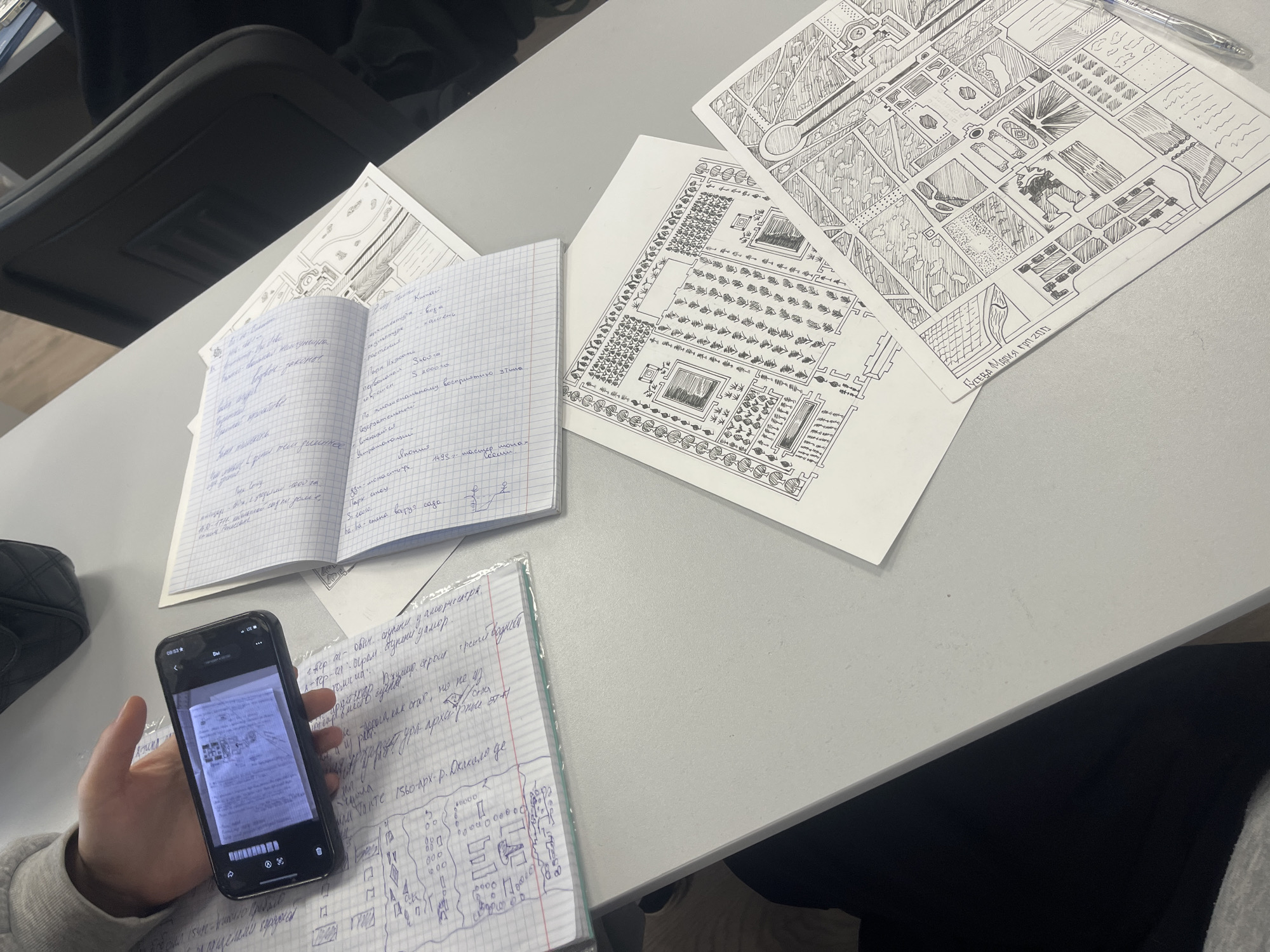 A top-down view reveals a table with several documents and sketches related to landscape architecture or garden design. A partially visible person is holding a smartphone, which is capturing an image of a notebook filled with handwritten notes. Several sketches of garden layouts are scattered on the table. Some notes are in Cyrillic script. In the upper right corner of one of the sketches, it reads “Гусева Мария гр. 2010”. This suggests the sketch was done by a person named Guseva Maria, possibly a student, in 2010. There is no information about Saint Petersburg in the image.
