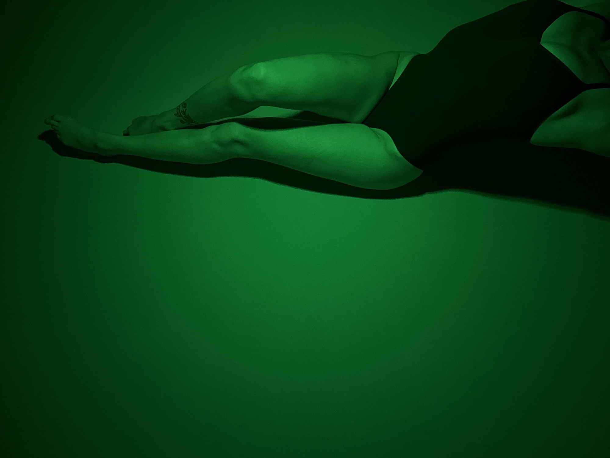 A person with visible legs, feet, and part of the torso, wearing dark bottoms, is lying on a solid green surface. The person's left leg (as seen by the viewer) is extended downwards while the right leg is bent at the knee. There is a tattoo visible on the ankle of the left leg. The scene is bathed in green light, creating strong shadows around the body. It is not possible to determine the location in Chaikovsky, Russia from this image.