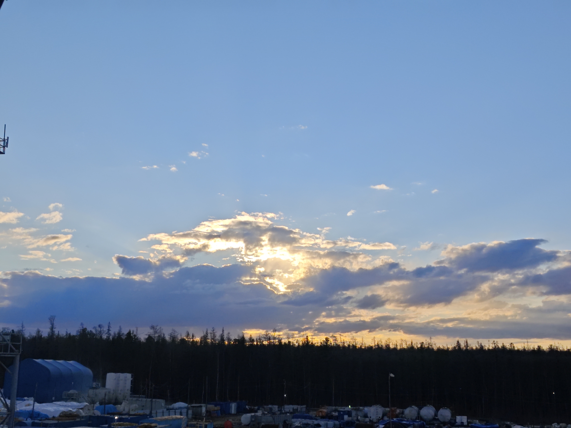 The image depicts a dusk scene in an industrial area near a forest. The sky is predominantly blue with scattered clouds, some of which are illuminated by the setting sun, creating a golden glow. Below the sky, a dense line of dark trees forms a horizon. In the foreground, there are various industrial objects, including large tanks, blue tents, and other unidentified equipment. The location is specified as Unknown, Russia. There are no people visible in the image.