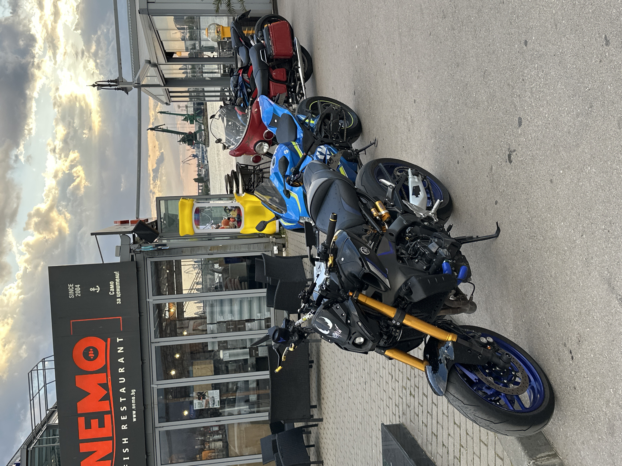 The image depicts a row of parked motorcycles in front of the "NEMO Fish Restaurant" in Varna, Bulgaria. The restaurant's sign is visible, indicating its establishment since 2004. The motorcycles consist of a black and blue Yamaha motorcycle in the foreground, followed by a blue Suzuki motorcycle, a red motorcycle, and a black motorcycle. The setting appears to be a waterfront area, with cranes and a harbor visible in the background. The sky is cloudy with a sunset or sunrise effect. Outdoor seating is present in front of the restaurant.