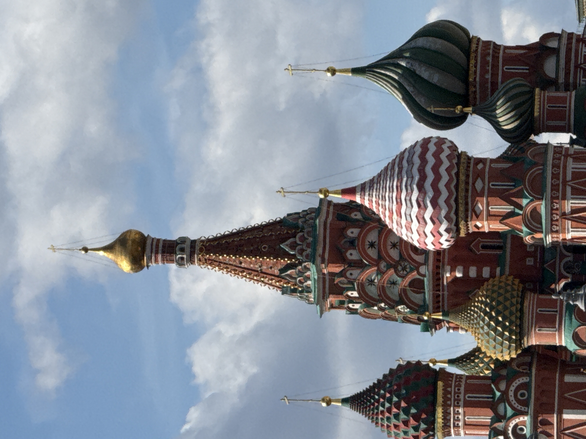 The image is a close-up, eye-level shot of the distinctive onion domes of Saint Basil's Cathedral, located on Red Square in Moscow, Russia. The sky is partly cloudy with blue patches visible. Multiple domes are in focus, showcasing their intricate architectural details: colorful patterns, gold finials topped with crosses, and ribbed textures. The domes vary in size and design, with some featuring red and white stripes, others green and gold, and one prominent central spire with a golden dome. The warm red and vibrant colors of the cathedral's facades are also visible, contrasting with the sky. No people or any actions are depicted in the frame.