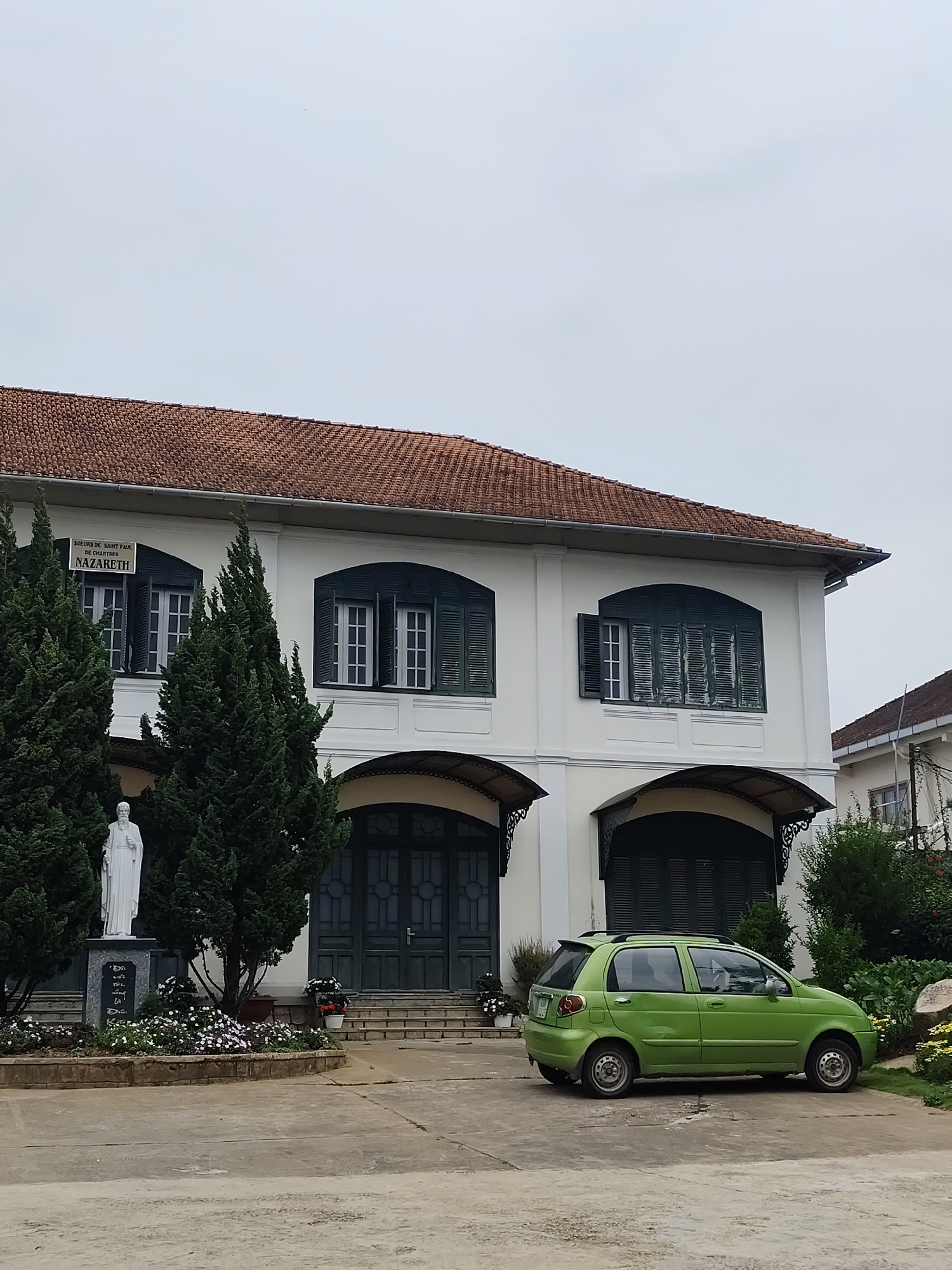 A daytime shot shows a white, two-story building with a red tile roof. The building has dark green shutters and a covered entrance. A statue stands to the left, surrounded by greenery and flowers. A small, bright green car is parked in front of the building. The building is identified by a sign as Soeurs de Saint Paul de Chartres Nazareth, which is located in Da Lat, Vietnam. The sky is overcast. The ground is concrete and appears to be a parking area or driveway.