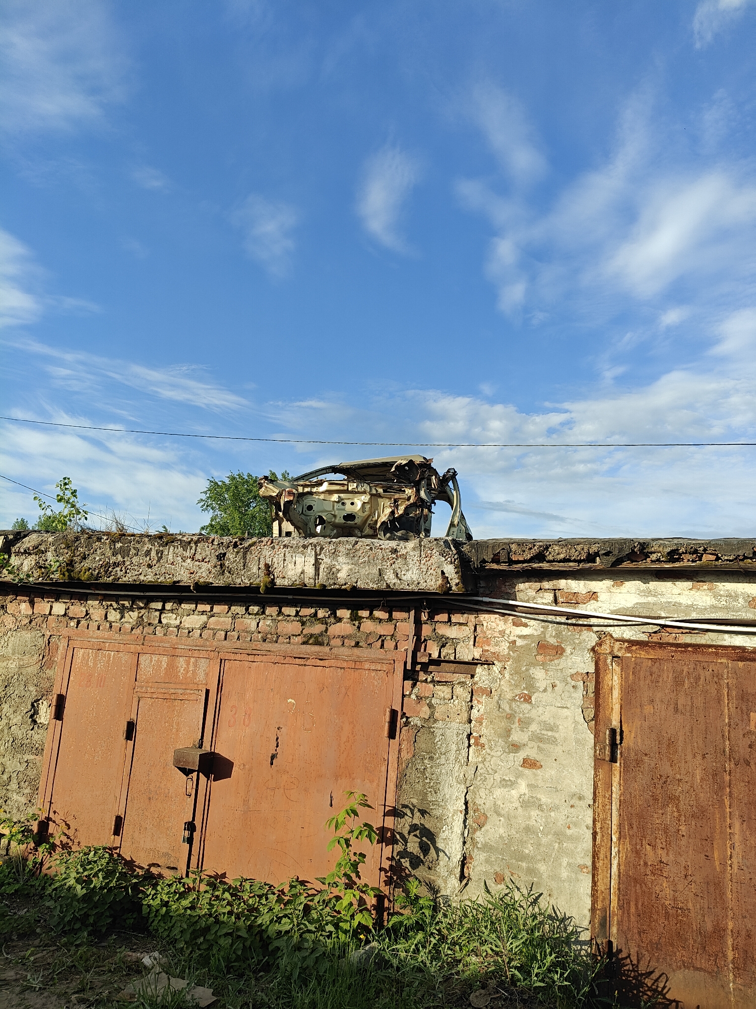 In Chaikovsky, Russia, the image depicts two rusty, red metal garage doors partially obscured by green weeds. Above the garage doors is a weathered brick wall topped with a concrete ledge. Atop the ledge sits the burned-out, skeletal remains of a car. The sky above is blue with scattered white clouds. A power line runs horizontally across the upper part of the scene.