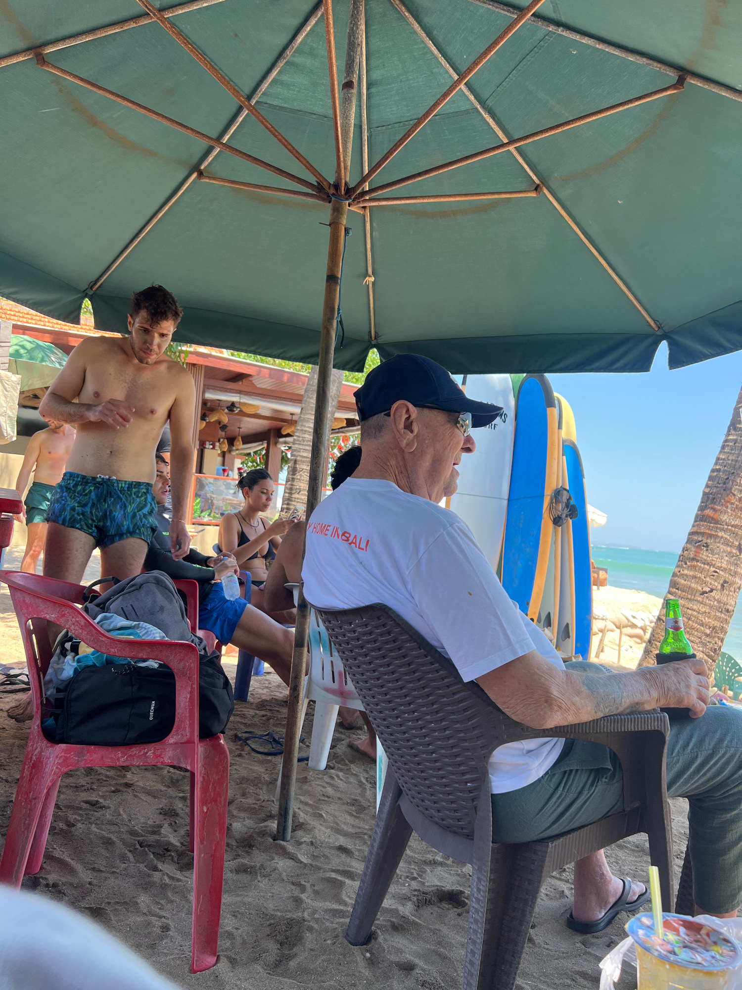 In Kuta, Indonesia, on a sunny beach, several people are gathered under a large green umbrella. In the foreground, an elderly man wearing a black baseball cap and sunglasses is seated in a brown plastic chair, holding a green bottle with a beverage. He is wearing a white t-shirt with orange text that reads "HOME IN BALI" and green pants. To his left, a young man with curly hair, shirtless and wearing patterned swim trunks, stands. Behind him, another man is visible in green shorts. A woman in a black bikini is seated to the right of the young man. Surfboards are propped up in the background, and the ocean is visible beyond the sandy beach. Several plastic chairs are scattered around, one red chair to the left holds a black backpack. The scene suggests a relaxed beachside gathering.