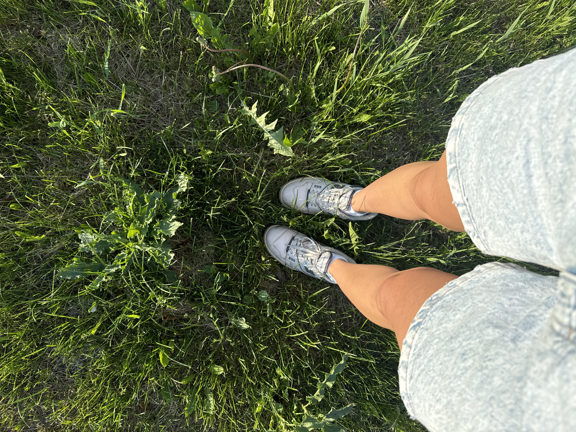 The image shows a first-person perspective looking down at a person standing in grassy terrain. The person is wearing light-colored denim shorts and white sneakers with "550" visible on one of the shoes. The ground is covered with green grass, some dry patches, and weeds. The location appears to be outdoors in a natural setting. The location is not specified as being in Bolshiye Vyazyomy, Russia.