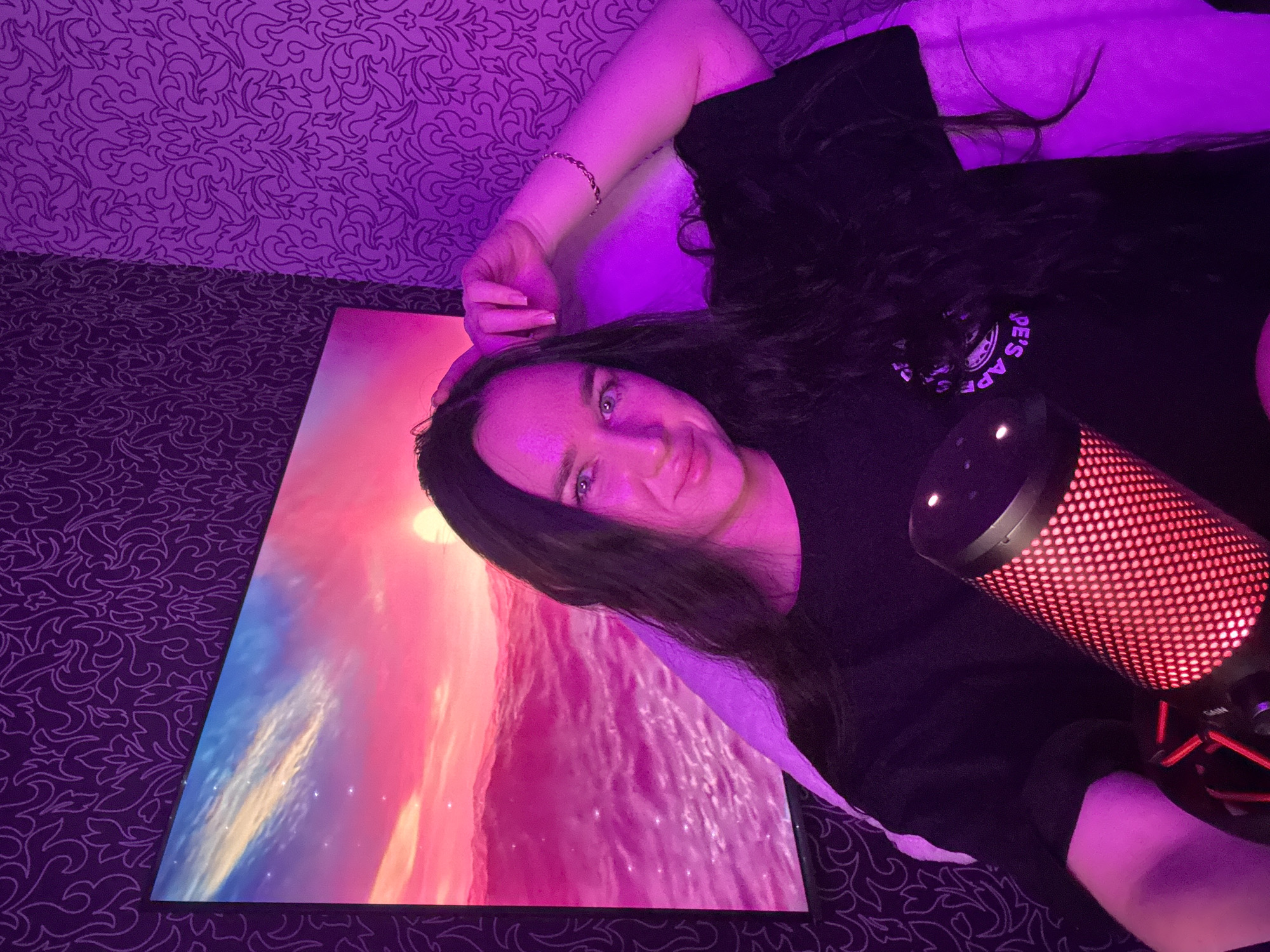 A young woman with long, dark hair is seated in front of a large screen displaying a vibrant sunset over a body of water. She is wearing a black t-shirt and has one hand resting on her head, while the other arm is bent at the elbow. In the foreground, a red microphone with a patterned grille is positioned on a stand. The background features patterned wallpaper illuminated by purple lighting. The scene appears to be a personal studio or gaming setup, potentially in an urban environment like Novosibirsk, Russia, given the context provided.