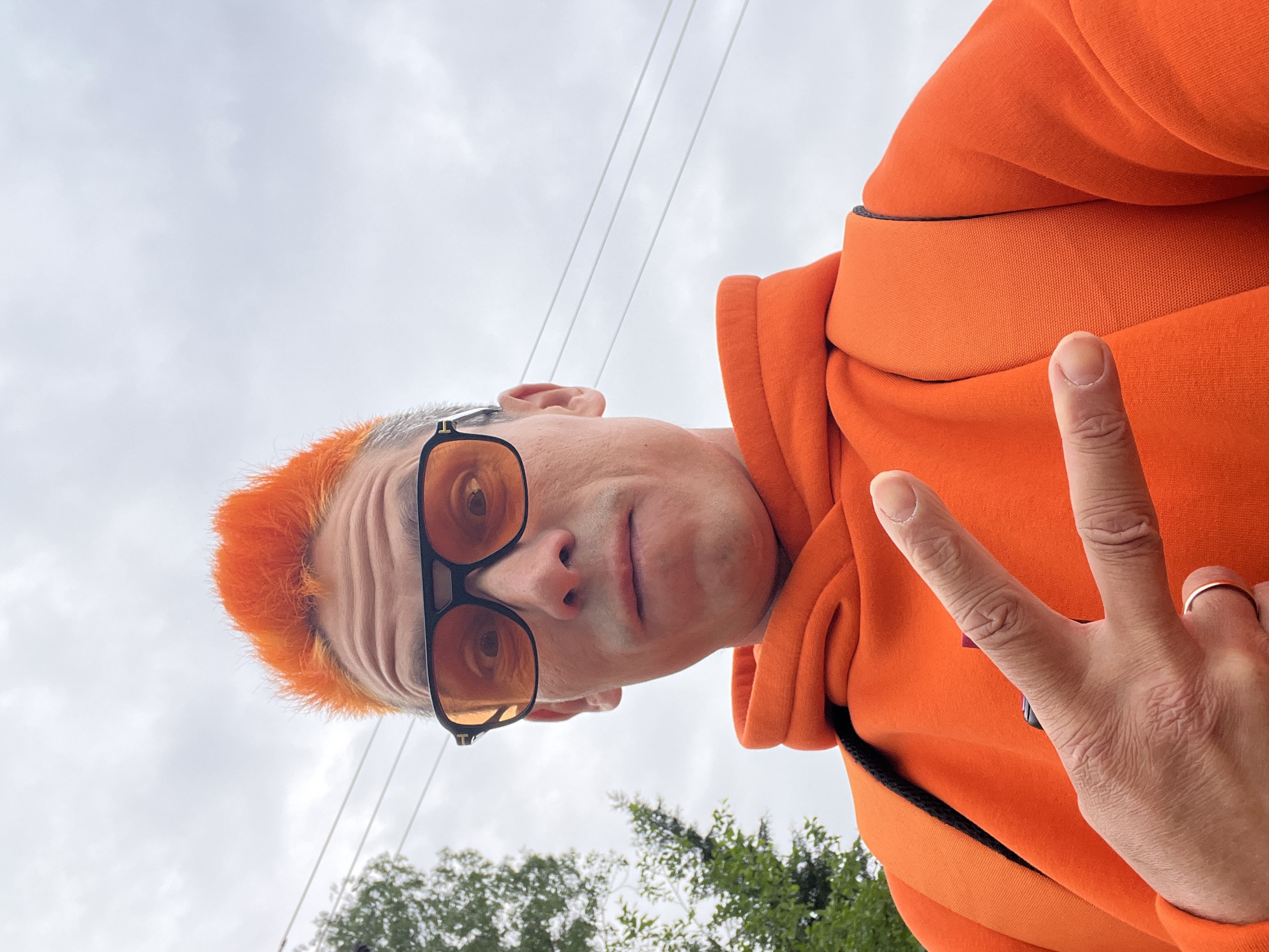 A selfie-style photograph features a man with orange hair and tinted glasses, wearing an orange hooded sweatshirt. He is holding up his hand in a peace sign. The background shows an overcast sky, power lines, and trees. It is implied that the location is Chaikovsky, Russia.