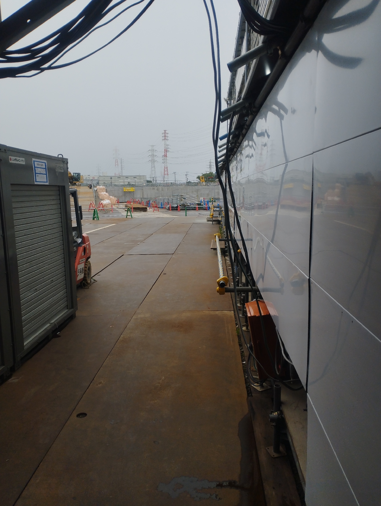 This is a photograph taken on a cloudy day at a construction site in Ichikawa, Japan. The scene is dominated by a large, reflective, grey metallic wall on the right, with scaffolding and electrical cables running along its side. To the left of the wall is a paved walkway. Further to the left, a grey, corrugated metal storage container is visible, with a red forklift parked next to it. In the background, there are stacks of sandbags, temporary fencing, and electrical pylons, indicating ongoing construction or infrastructure work. The ground is paved and appears wet in places. The overall impression is of an industrial or utility-focused area within the city.