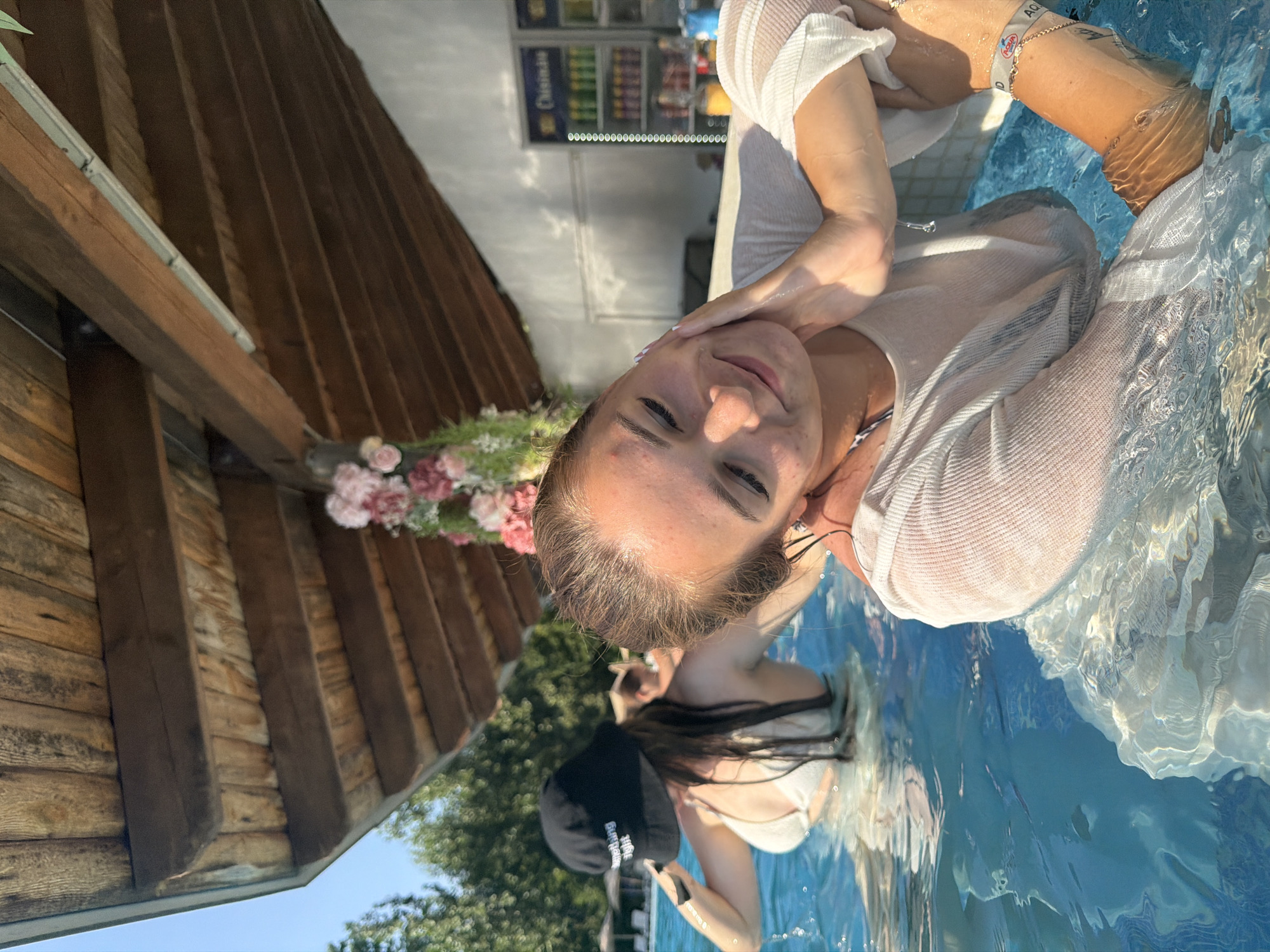 The image shows two women in a swimming pool, likely at a resort or hotel in Sociteni, Moldova. The woman in the foreground is leaning forward, her face illuminated by the sun, with her eyes closed and a gentle smile. She is wearing a wet, sheer white top that clings to her body. Her right arm is extended, and a bracelet with the word "Aqua" is visible on her wrist. The woman in the background, to the left, is wearing a white bikini top and a black bucket hat with "Something Right" printed on it. She is looking over her left shoulder, her hands in the water. The pool water is a vibrant blue. Above the pool, there is a wooden-beamed roof structure adorned with pink and white flowers. In the background, a white wall is visible, with refrigerators containing drinks. Trees and lounge chairs are also visible in the distance, suggesting an outdoor setting. The scene captures a moment of relaxation and enjoyment in a warm climate.