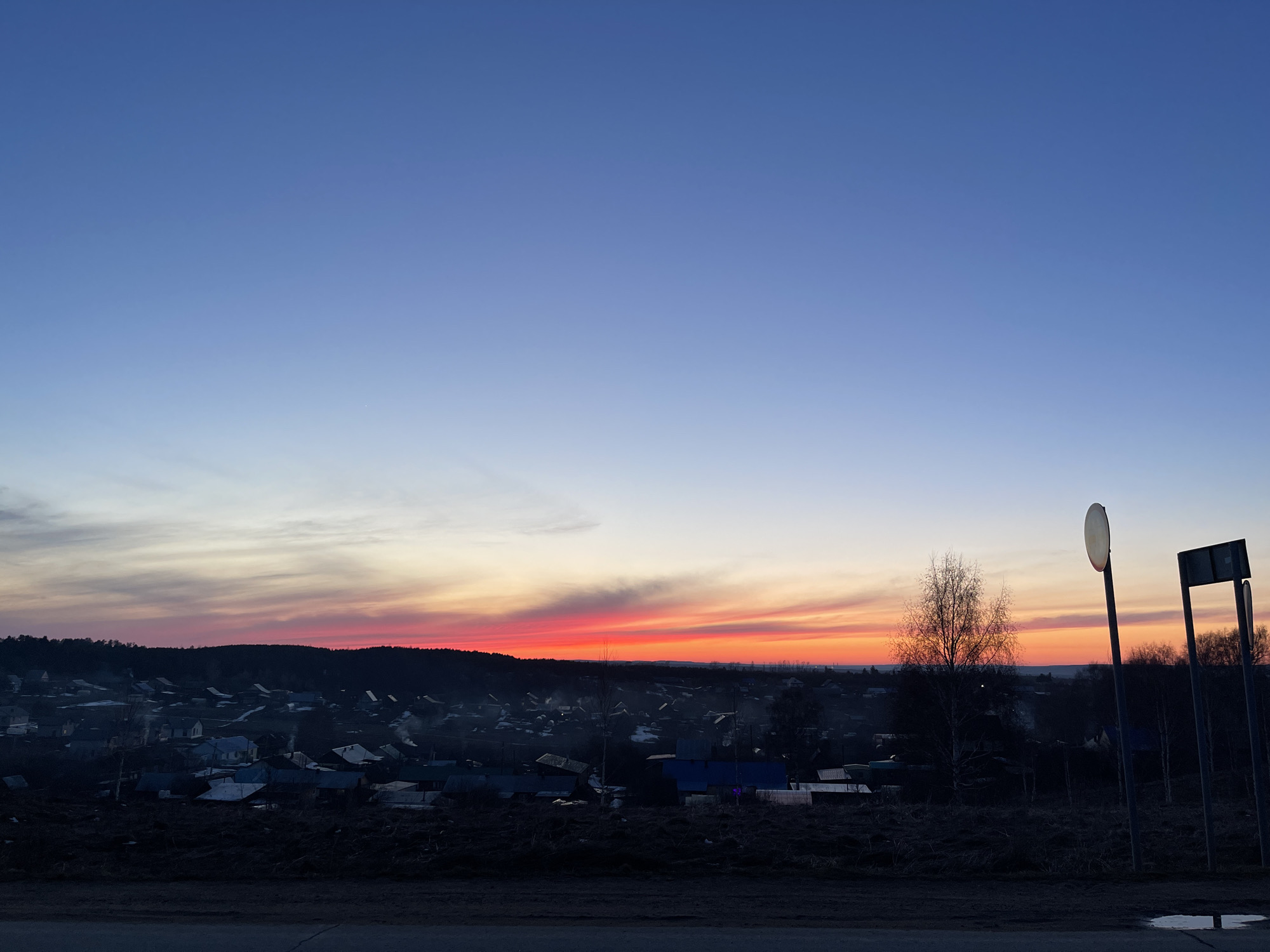 The image depicts a rural landscape at twilight in Фоки, Russia. The sky transitions from deep blue above to a vibrant red and orange along the horizon, indicating either sunrise or sunset. Below the horizon, there is a silhouette of a densely packed village with numerous houses. The foreground features a dark field or vegetation-covered area and a section of road in the very bottom of the frame. In the right foreground, several weathered roadside signs stand, casting long shadows. The scene is quiet and still, suggesting a peaceful rural setting.