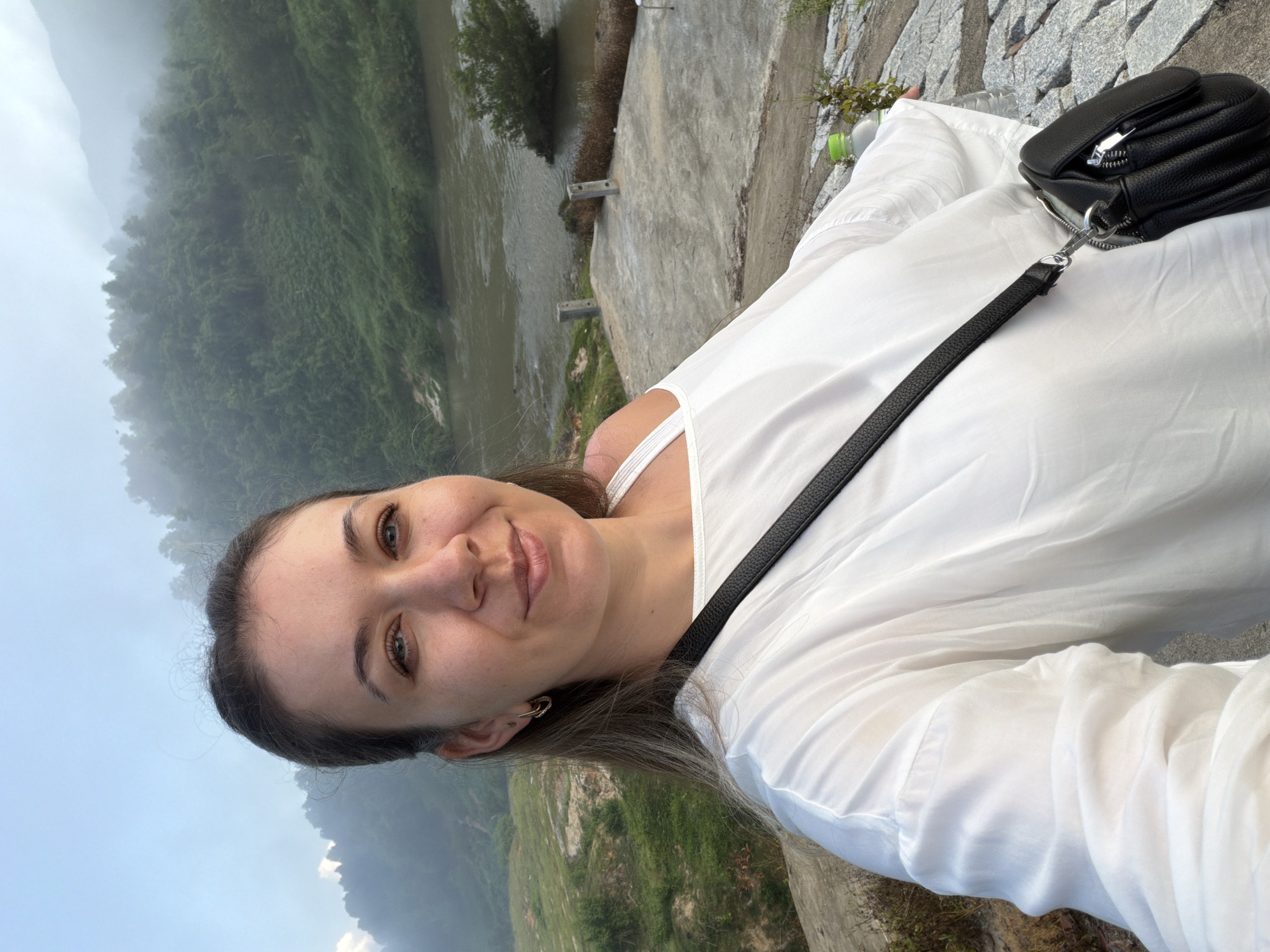 A woman with long brown hair, wearing a white top and a black crossbody bag, is taking a selfie outdoors. She is smiling and looking at the camera. Behind her, a hazy landscape of green hills and a river is visible. The scene appears to be in the city of Nam Khanh Vinh, Vietnam, suggested by the dense vegetation and the style of the surrounding terrain. The ground in the foreground is made of stone and concrete. A plastic bottle of water is also visible to the right of the woman.