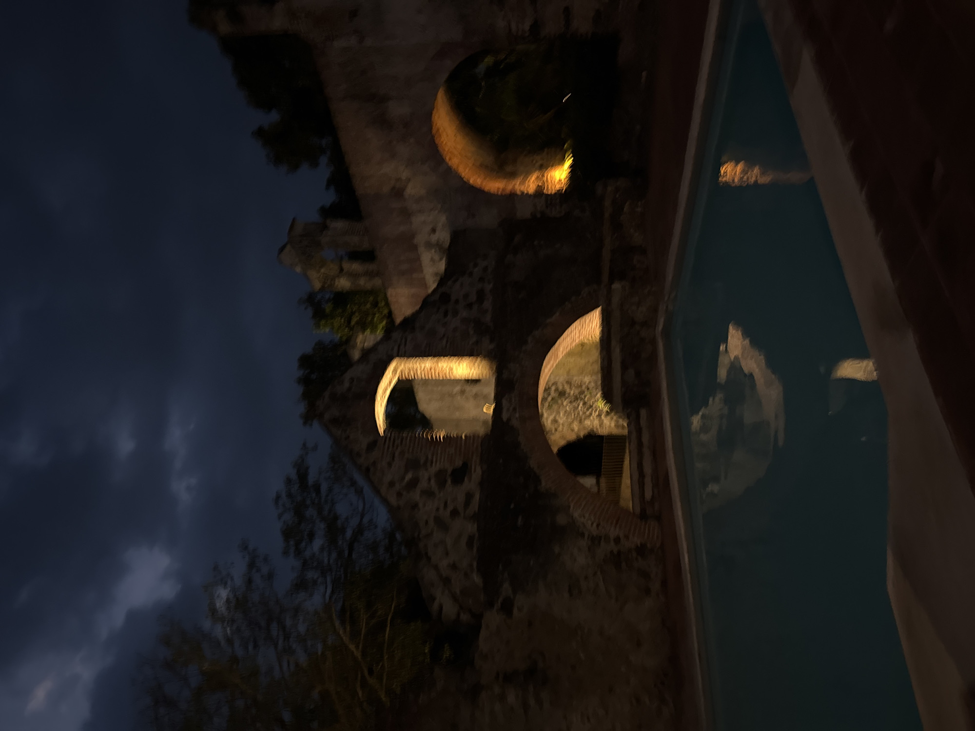 The image captures the Hacienda de Cocoyoc in Cocoyoc, Mexico, at night. The illuminated stone arches of the old aqueduct or building ruins are a prominent feature. The sky is visible in the background, hinting at nightfall. A body of water, possibly a pool, reflects the structures, adding a sense of depth to the scene. The lighting is warm and highlights the architectural details. Trees are present in the foreground, contributing to the overall ambiance. There are no people visible in the shot.