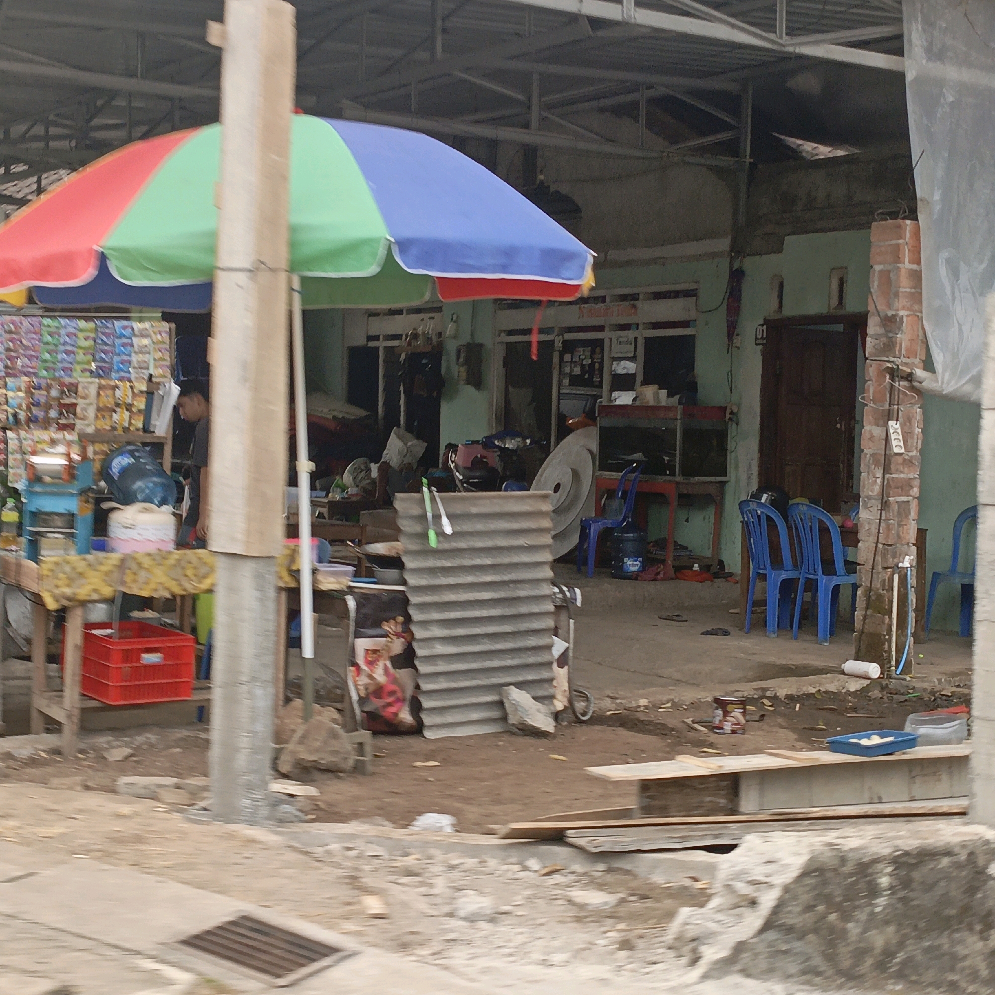 This image depicts a street-level view of a shop or stall in Salatiga, Indonesia. A prominent, multi-colored umbrella, with sections of red, green, and blue, is set up, partially obscuring the view of the establishment behind it. To the left, a rack of packaged goods, possibly snacks or toiletries, is visible. In front of this rack, a blue water dispenser with a large plastic bottle is situated. A person, appearing to be a male, is standing behind the counter, facing away from the camera. Further back, there are chairs and tables, and a doorway is visible in the building. The ground appears to be dirt and concrete. Construction materials, including corrugated metal sheets and bricks, are present, suggesting ongoing renovation or a rustic building style. A red plastic crate is placed on a wooden table in the foreground. In the background, other structures and the general environment of a local street or market are discernible.