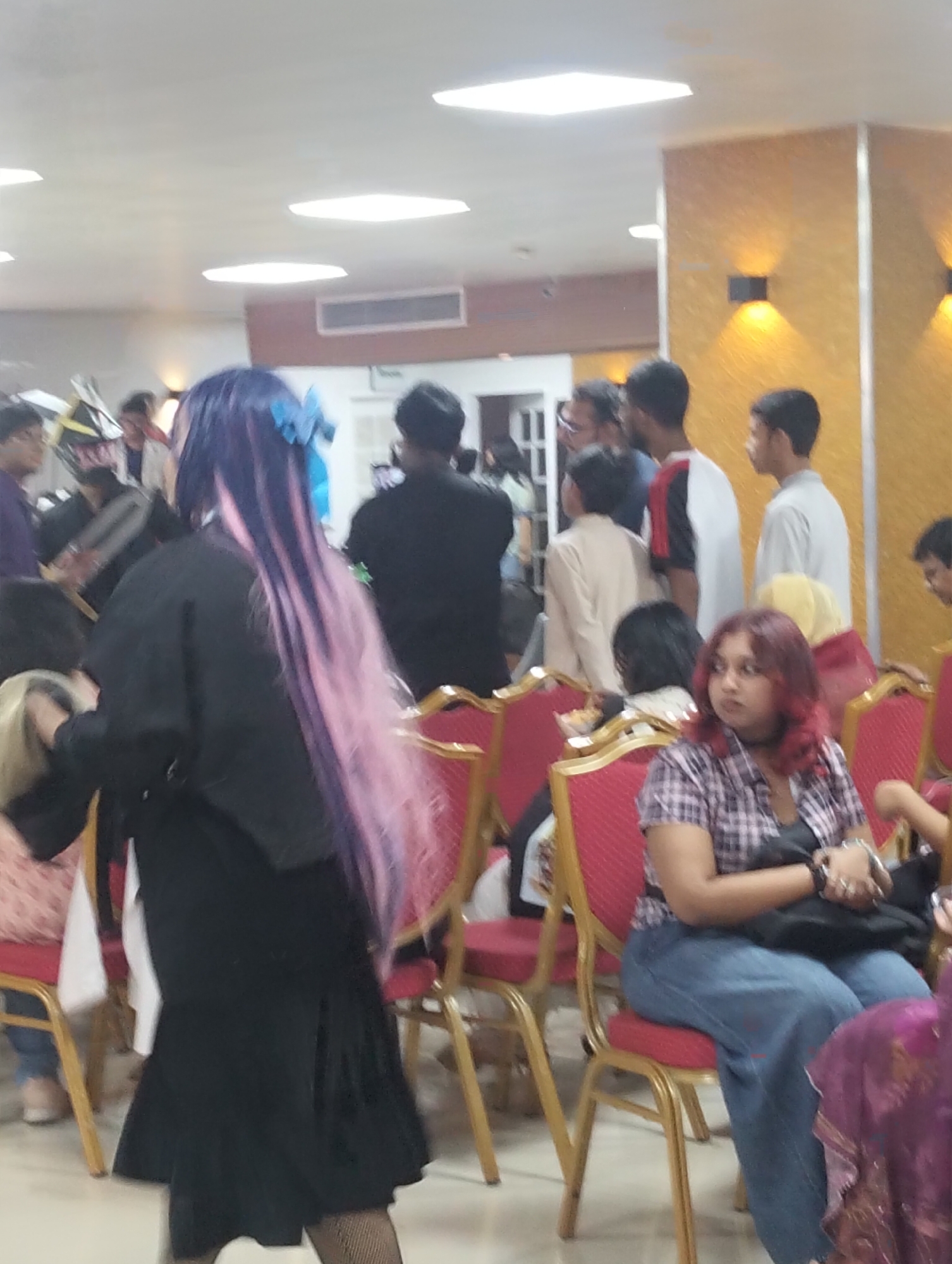 In a brightly lit indoor space with a wood-paneled wall and white ceiling lights, a group of people are gathered. The scene appears to be an indoor event or convention. In the foreground, a person with long, purple and pink hair is seen from behind, wearing a black outfit. To the right, a woman with red hair is seated on a red chair, wearing a plaid shirt and blue jeans. Other individuals are visible in the background, some standing and others seated, in various casual attire. The presence of multiple people in an indoor setting suggests a public gathering or event. The image does not provide specific visual cues to pinpoint the exact location within Dhaka, Bangladesh.