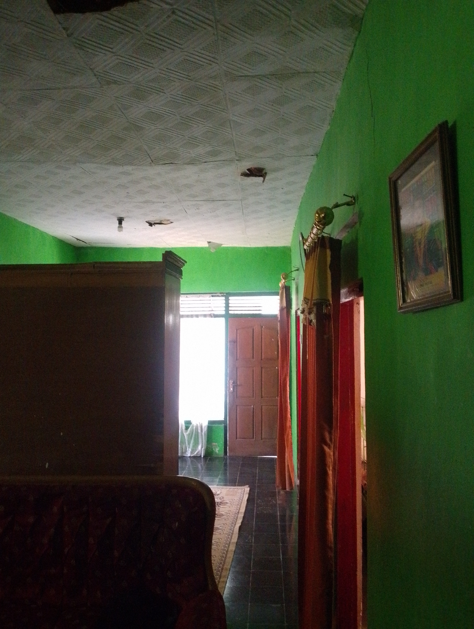 The image depicts the interior of a room, likely a hallway or living area, in a residential building located in Sukamanah, Indonesia. The walls are painted a vibrant green. The ceiling is made of patterned tiles and shows signs of wear, with some visible cracks and damage. A single light bulb hangs from the ceiling, illuminating the space.
On the left side of the frame, a large, dark brown wooden cabinet or wardrobe dominates the scene. In the foreground, the top of a dark red, upholstered sofa is visible. A patterned rug is laid out on the dark tiled floor, extending towards a doorway.
The doorway, on the right side of the hallway, features a wooden door with multiple panels and is open, allowing light to stream in. Beyond the door, a window with horizontal blinds is visible, and sheer white curtains hang. Two sets of curtains, one in a reddish-brown hue and another in a lighter brown, are draped from rods on either side of the doorway, partially obscuring the view further inside.
On the green wall to the right, a framed picture hangs. The frame appears to be made of dark wood, and the picture within depicts a landscape or still life. A decorative metallic lamp with a golden finish is mounted on the wall above the curtains. The overall impression is of a simple, lived-in home.