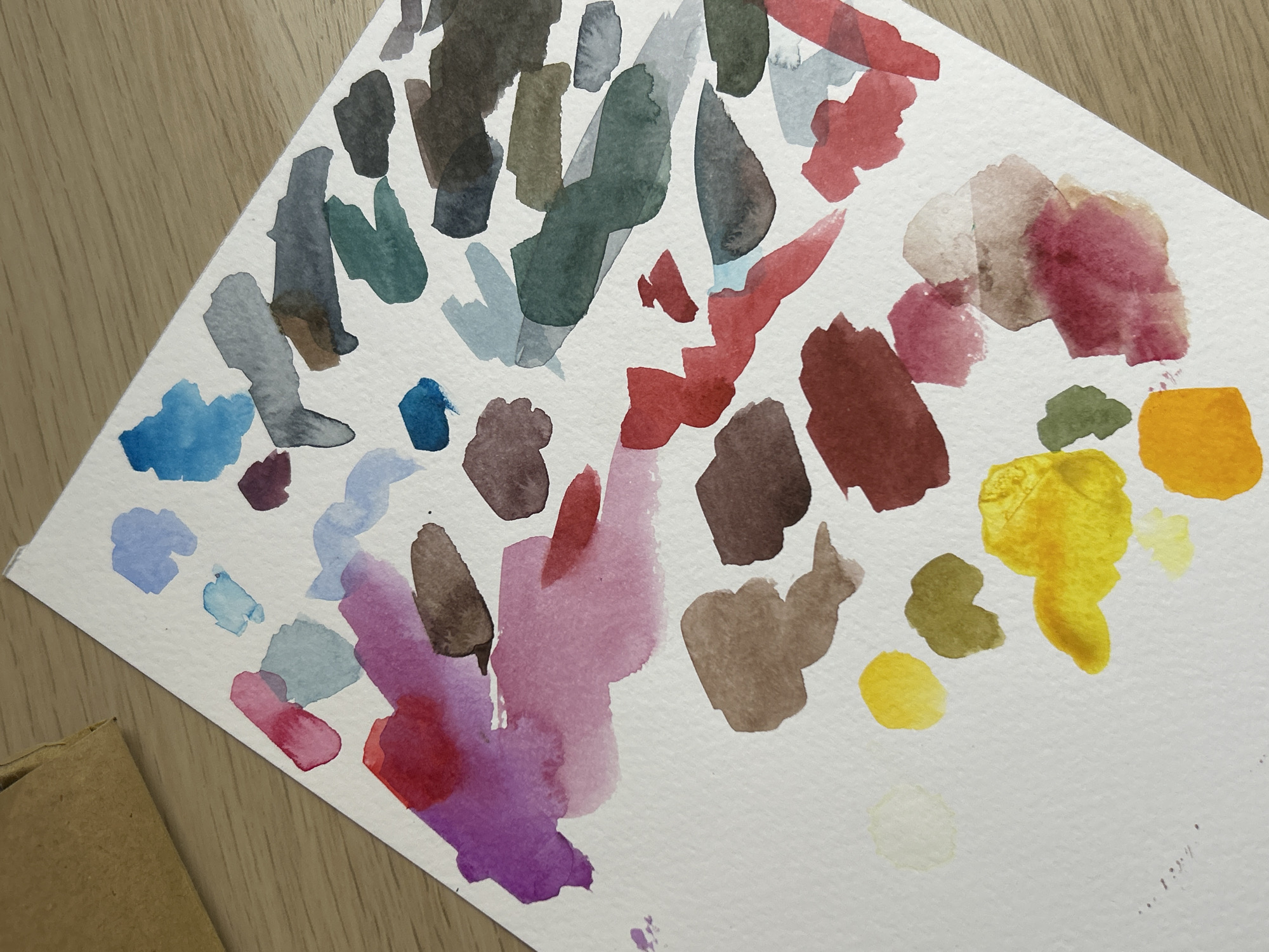 A top-down view of a rectangular white piece of paper on a light wood surface. The paper is covered with irregularly shaped swatches of watercolor paint in various colors including blues, grays, greens, reds, purples, browns, and yellows. Some of the colors blend into each other. Part of a brown cardboard object is visible in the lower left corner of the image.
The location in the city of Sofia, Bulgaria is not discernible from the image.