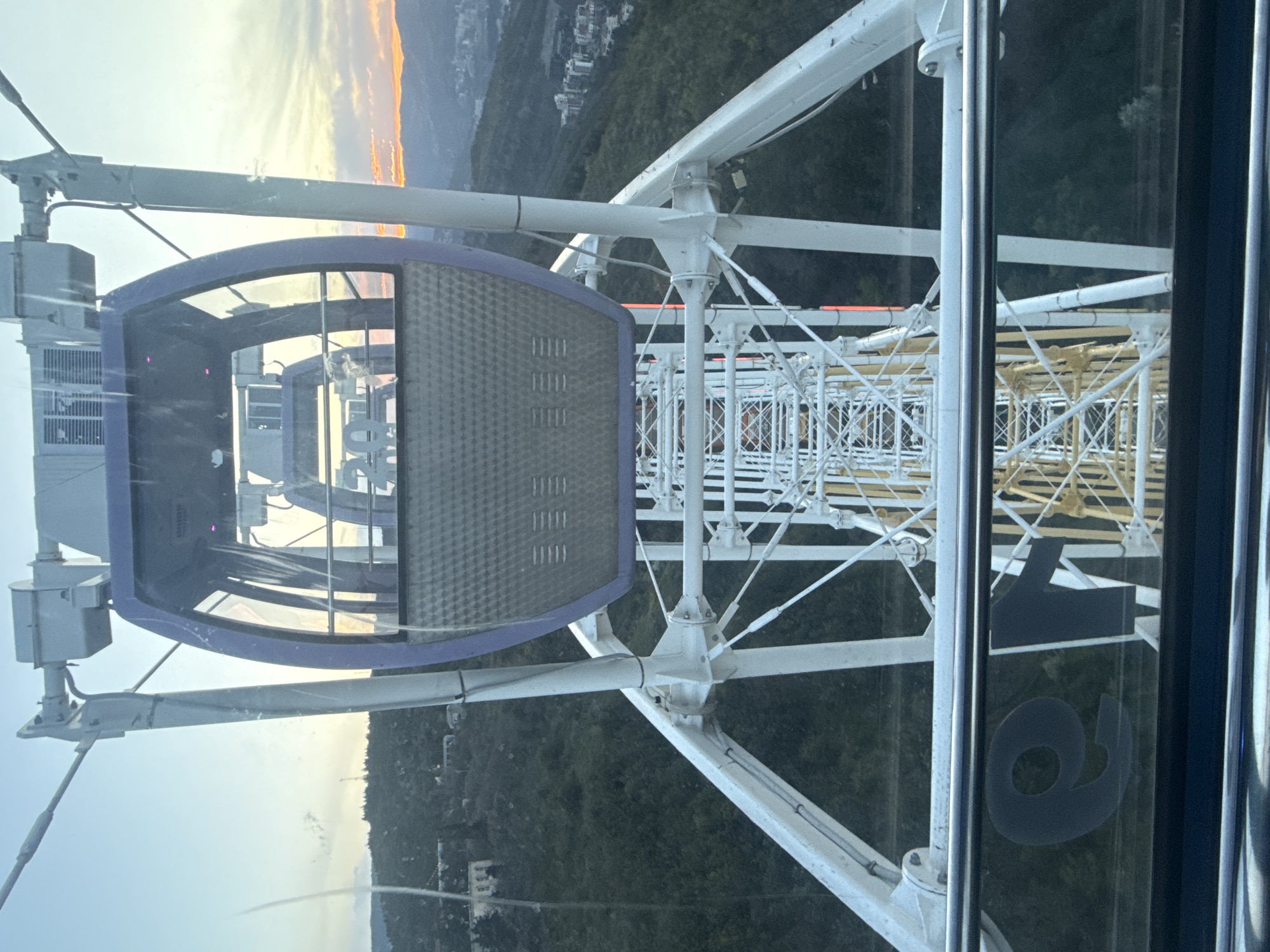 The image shows a view from inside a Ferris wheel cabin, likely in Tbilisi, Georgia, as the Tbilisi Ferris wheel is a well-known attraction. The cabin has a blue frame and a gray mesh panel. The number "19" is visible on a glass panel, likely the cabin number. The Ferris wheel's white metal structure extends upwards and downwards from the cabin. Outside the cabin, there is a view of a city, mountains, and a sunset. Green vegetation can be seen on the mountains.