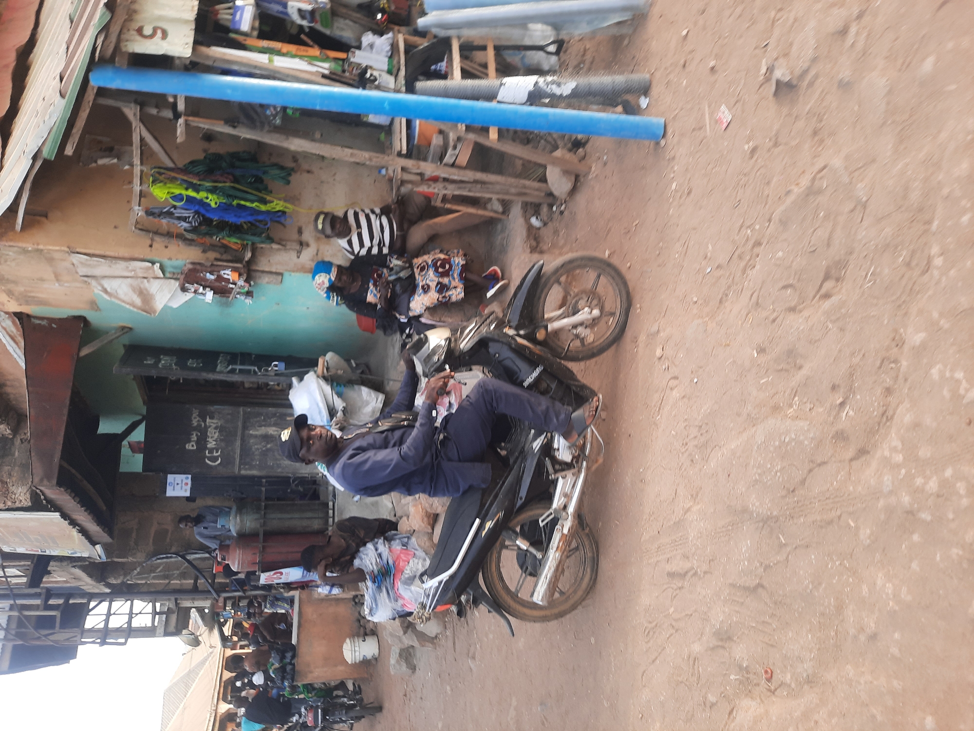 The image shows a street scene in Gwong, Nigeria. A man is sitting on a black motorcycle, casually dressed in a light blue outfit and cap, and appears to be looking directly at the camera. Behind him, two people are seated, one in a striped shirt and the other in a patterned dress with a blue head covering. A storefront is visible in the background, displaying items such as colorful ropes, pipes, and building materials. A sign that reads "Buy your Cement" is visible as well. In the lower part of the picture, more motorcycles are parked. The ground is dirt, and the scene appears to be in a commercial or market area.