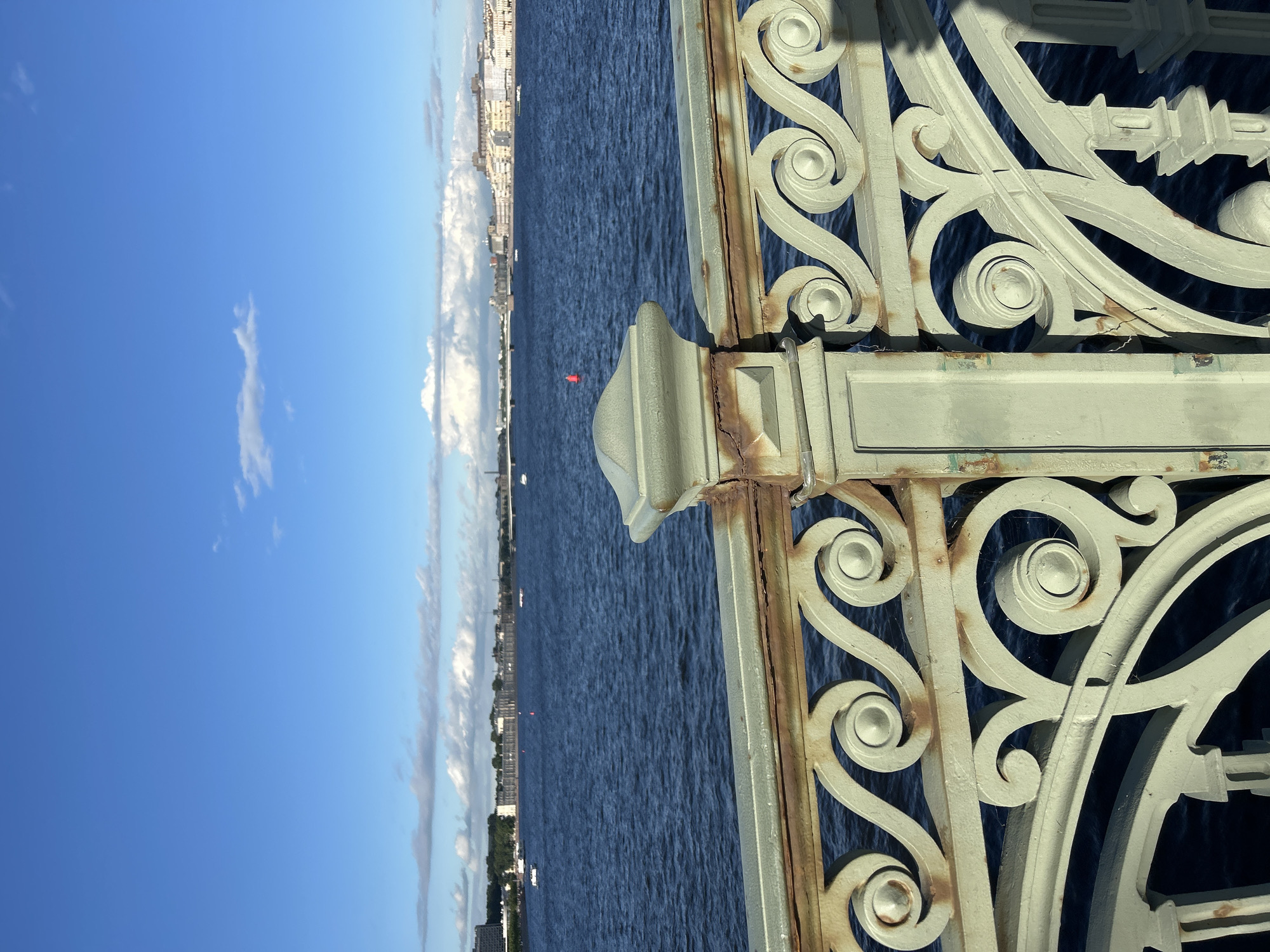 An ornate, light green, metal bridge railing with visible rust spots runs across the foreground of the image, obscuring the lower portion of the scene. Behind the railing, a wide body of deep blue water stretches into the distance. The water is calm with slight ripples. Several small white boats are scattered across the water. On the far side of the water, a city skyline is visible, characterized by a line of buildings and structures stretching across the horizon under a bright blue sky with scattered white clouds. A red buoy is present in the water closer to the foreground. The image appears to be taken from a bridge overlooking a river or bay.