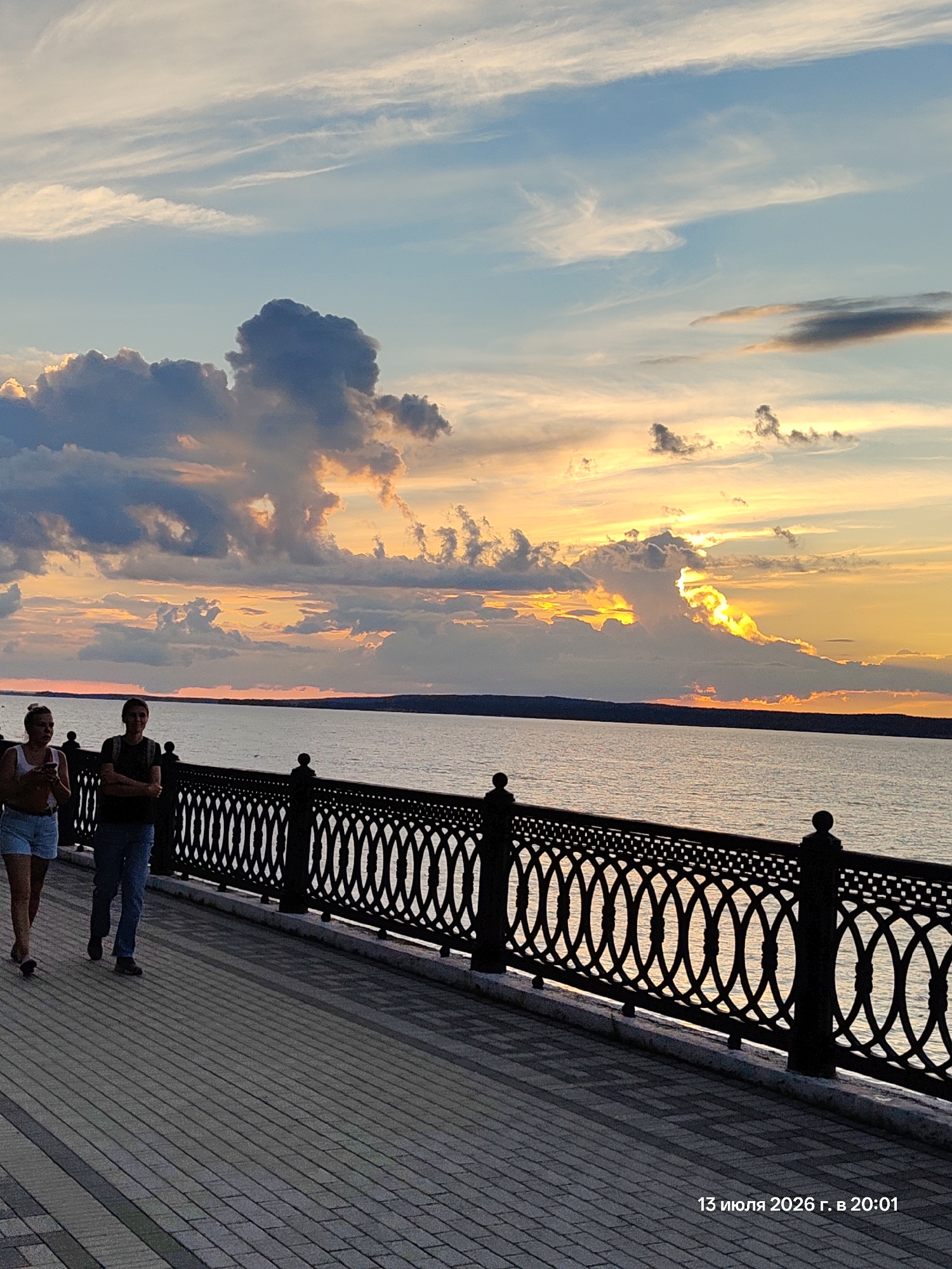 A sunset over a body of water is depicted, with a man and a woman walking on a paved promenade beside a decorative metal railing. The sky is filled with dramatic clouds, illuminated by the setting sun. The water reflects the warm colors of the sunset. The promenade appears to be made of rectangular paving stones. The image is dated "13 июля 2026 г. в 20:01".