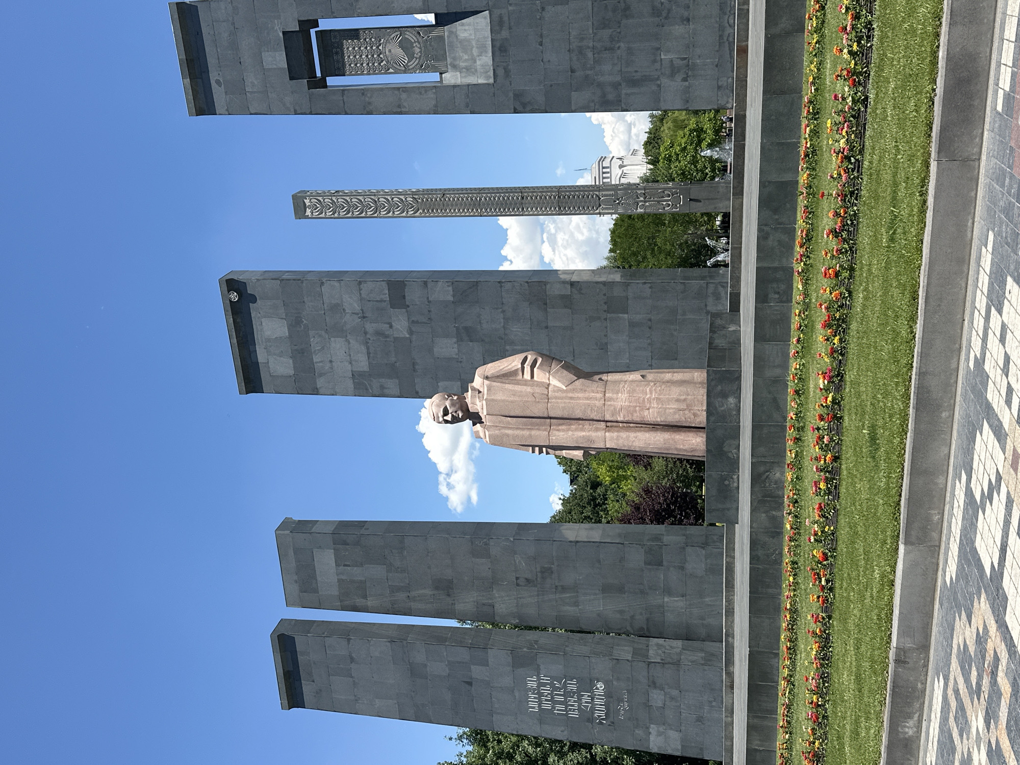 An outdoor monument scene is captured under a bright, clear blue sky with scattered white clouds. The central subject is a full-body statue of a man, rendered in a reddish-brown material, possibly bronze or stone, and depicted wearing a long coat or uniform. He stands upright between several imposing, tall, dark grey rectangular steles. These steles are constructed from large, squared blocks of stone.
Two steles flank the statue directly, with additional steles visible behind and above. One stele, in the upper right, features an intricate, carved, open design near its top. Another, positioned centrally behind the statue, is a slender, vertical column adorned with ornate patterns along its length. On a stele to the lower left of the statue, Armenian text is clearly visible, reading: "ՄԻԱՑՅԱԼ ՄԱՔՐԱՇԱՐԺ ԱՆՄԱՀ ՀՈԳԻՆԵՐԻՆ" which translates to "TO THE UNITED IMMORTAL SOULS."
In front of the statue and steles, a neatly maintained strip of vibrant green grass stretches across the frame, bordered by a long, narrow flower bed showcasing colorful blooms in shades of red, orange, and yellow. To the far right, a section of patterned pavement with white and dark grey geometric tiles is visible. In the background, beyond the monument, lush green trees are seen, and a glimpse of a distant white building with classical architectural elements suggests a city setting. The scene evokes a solemn and well-kept public memorial or park, likely captured during daytime in Yerevan, Armenia.