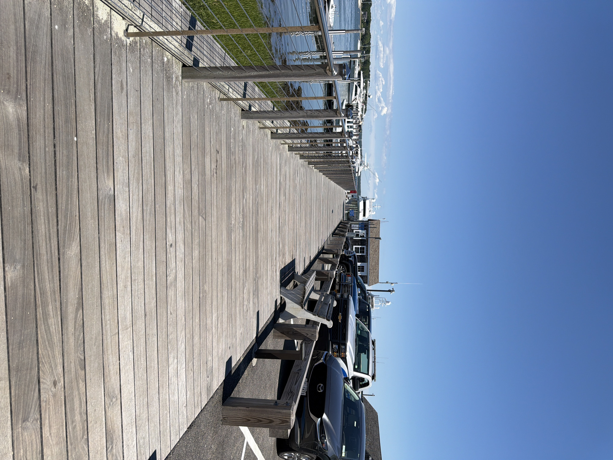 The image captures a wooden boardwalk extending into a harbor on a sunny day in the Village of Sag Harbor, United States of America. The boardwalk, made of weathered gray planks, is bordered on the left by a metal railing and lush green grass, and on the right by a parking area. Several boats, including white yachts, are docked in the calm, blue water to the left of the boardwalk. In the background, a few buildings, including a white structure resembling a small house and another with a dark roof, are visible. Parked cars, including a dark SUV, a black pickup truck, and a white truck with emergency lights, are situated along the right side of the boardwalk. A single, empty wooden bench is placed on the boardwalk, facing the water. The sky above is a clear, bright blue, with no clouds visible.