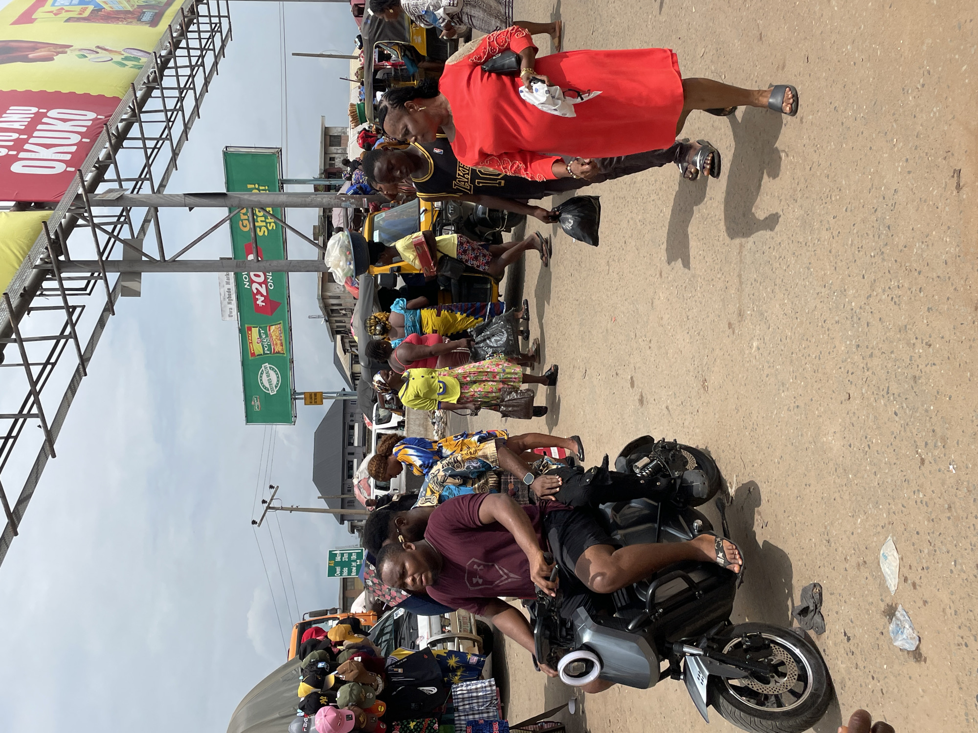 The image shows a street scene in Onitsha, Nigeria. A man sits on a motorcycle parked on the right side of the road, near a vendor selling hats. Other people are standing and walking nearby, including a woman in a red dress. Street signs and advertising billboards are visible in the background.