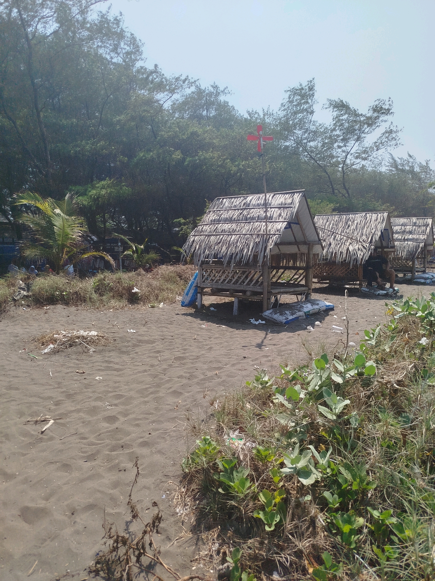 The image depicts a beachfront scene in Kenoyajaya, Indonesia. In the foreground, dark, coarse sand is visible with scattered debris, including plastic bottles and dried vegetation. To the right, lush green ground vegetation grows in clumps amongst dry grasses.
The midground features a row of three small, rustic huts with thatched roofs constructed from dried palm leaves. These huts are elevated on wooden posts and appear to be made from bamboo. The first hut, closest to the viewer, has a blue and white striped bag placed on the ground in front of it. The second hut has a person seated inside, wearing dark clothing. The third hut is partially visible.
In the background, a dense line of trees with dark green foliage borders the beach. A red, cross-shaped object on a pole is visible above the first hut, silhouetted against a pale blue sky. Some palm trees are also present on the left side of the image, near the trees. The overall environment suggests a natural coastal setting with signs of human use and some disarray due to debris.