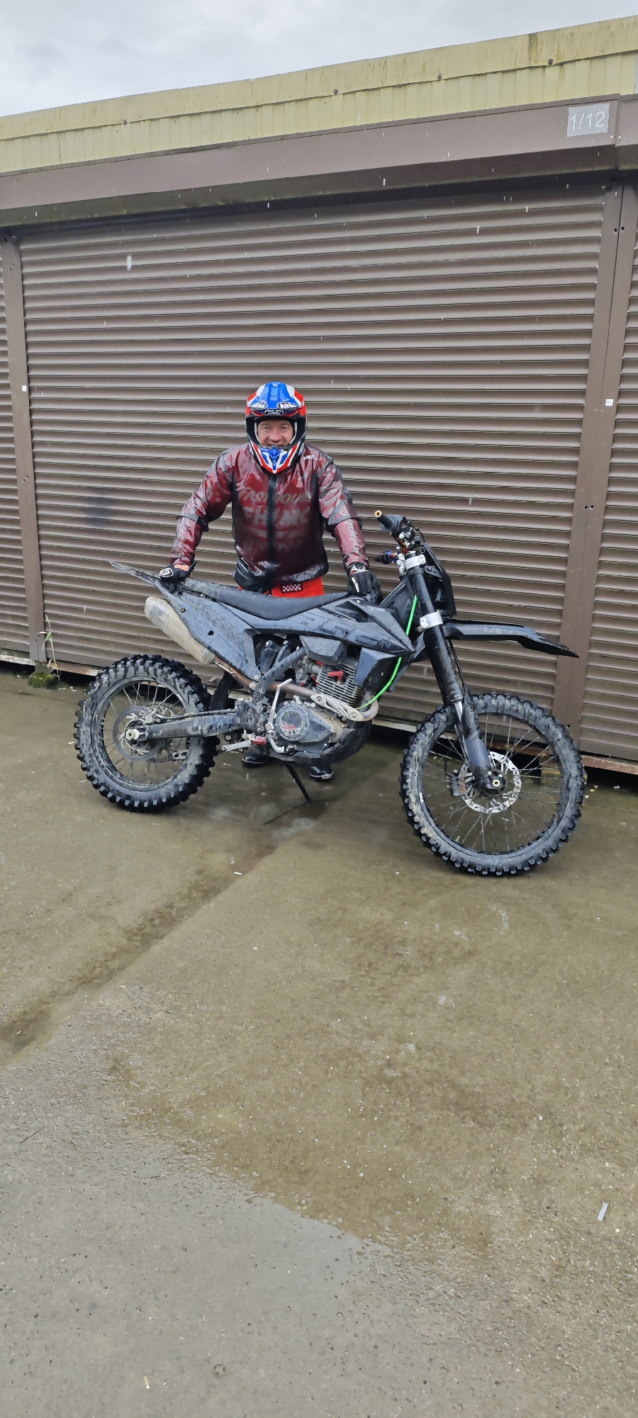 A man wearing a blue, white, and red helmet and a red jacket is posing next to a black dirt bike on a wet, gray concrete surface. The background is a brown corrugated metal wall with the number "1/12" visible in the top right corner. It is raining. The tires of the dirt bike are muddy. Based on the presence of water and dirt, the environment is likely outdoors. The location is Kazachiy Brod, Russia.
