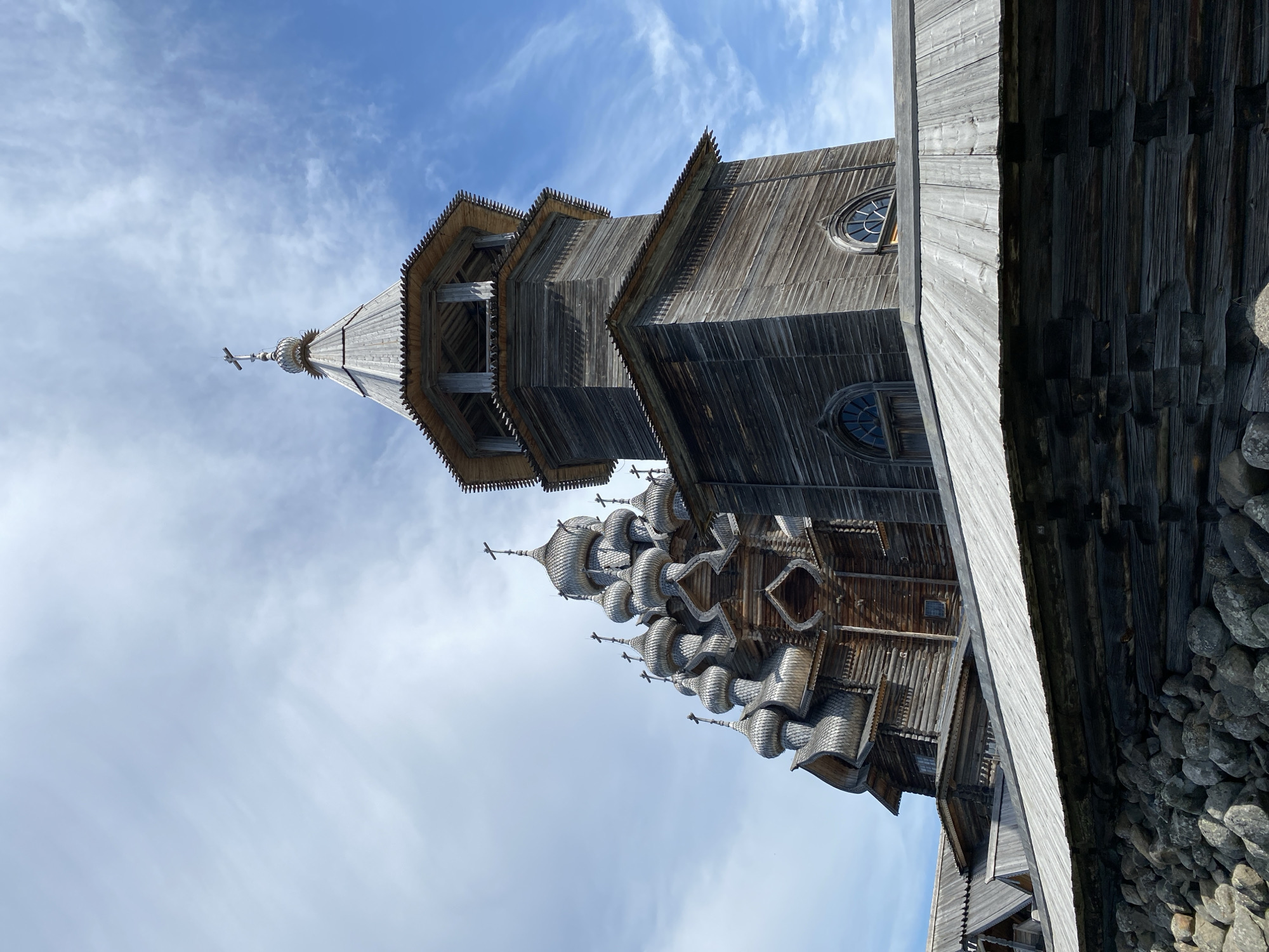 The image shows the Church of the Transfiguration, a wooden Orthodox church located on Kizhi Island in the Republic of Karelia, Russia, near the city of Boyarshchina. The church is constructed entirely of wood, with a multi-domed roof and intricate architectural details. In the foreground, a stone wall made of large, rough-hewn rocks separates the viewer from the wooden structures. Above the wall, the lower portions of the church complex are visible, featuring layered wooden roofs and walls made of logs. A prominent bell tower, also constructed of wood, rises on the right side of the image, topped with a conical roof and a cross. The main body of the church, visible to the left, is characterized by its numerous smaller domes, each adorned with a cross. The sky above is a vibrant blue with scattered white clouds. There are no people visible in the image.