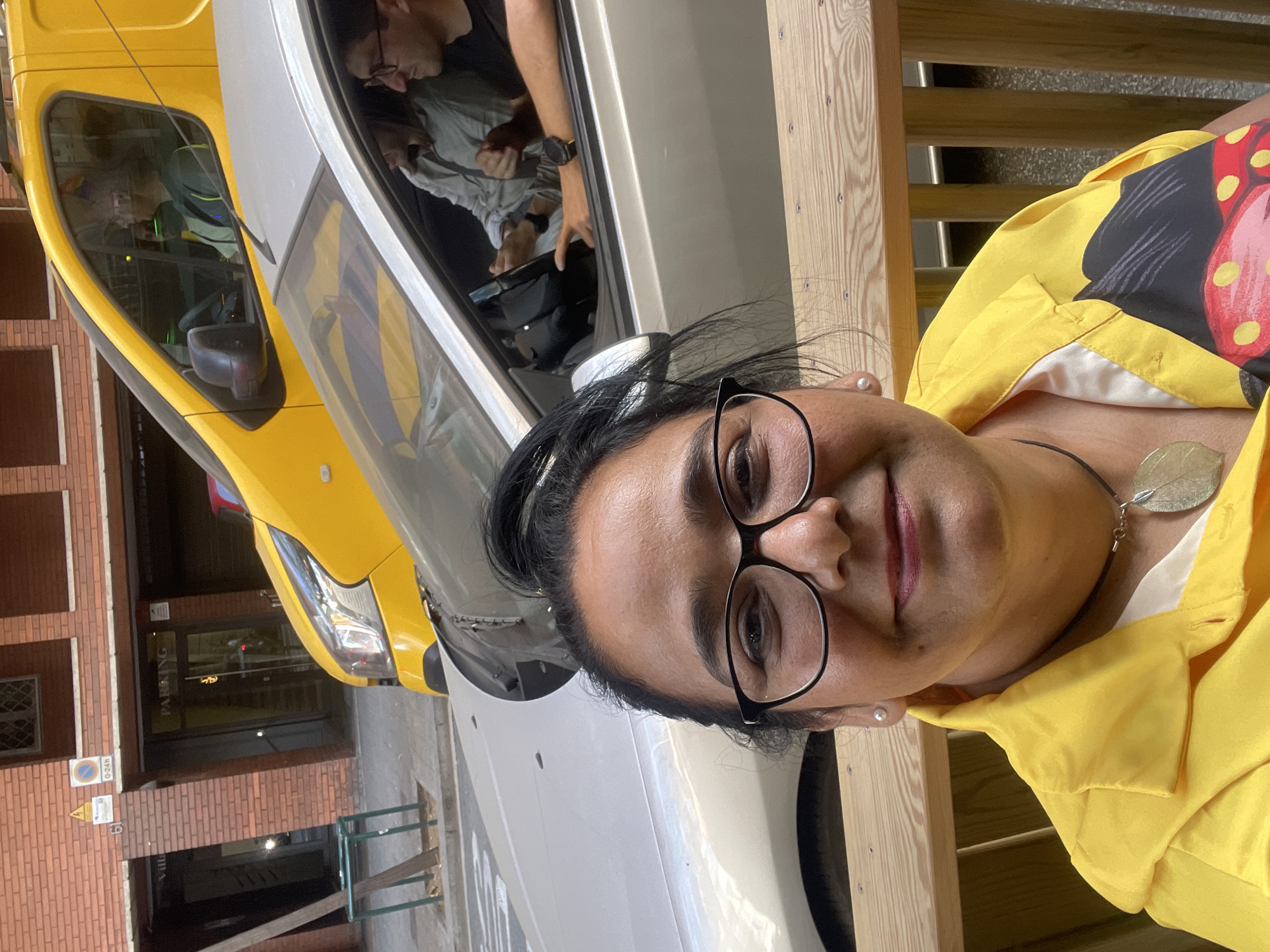 In Barcelona, Spain, a woman with dark hair pulled back and wearing glasses and a bright yellow shirt with a pattern is taking a selfie. She is seated outdoors on a wooden bench with a wooden railing behind her. In the background, two cars are visible: a silver car in the foreground with two people inside, and a yellow van behind it, also with a person in the driver's seat. The street appears to be lined with brick buildings. One building has signage indicating "PARKING" and another has "Villa."