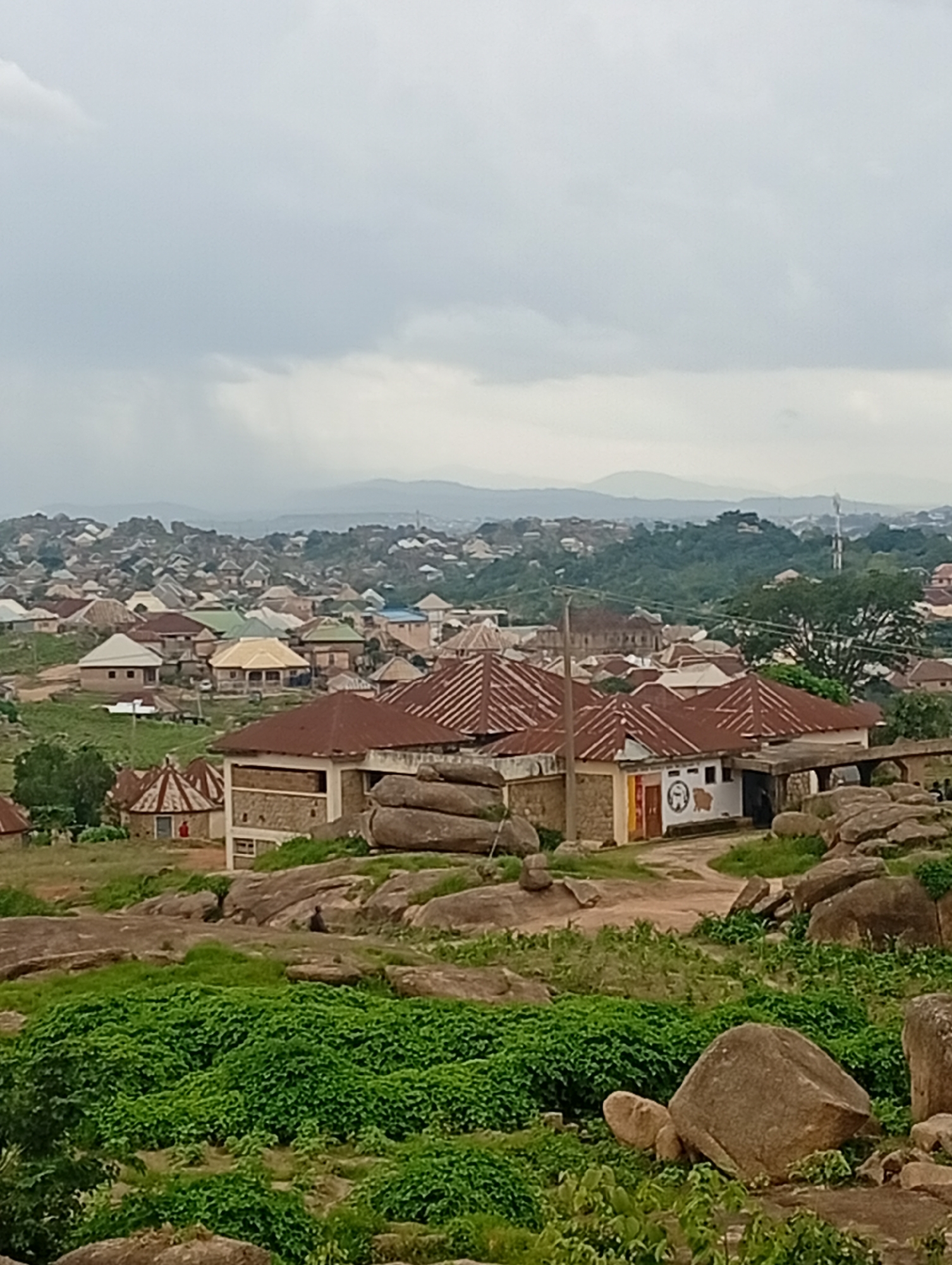 This is a high-angle, medium shot of a residential area on a rocky terrain in Jos, Nigeria. The foreground is dominated by large, weathered boulders and lush green vegetation. In the midground, several buildings with corrugated metal roofs are visible. These structures appear to be made of stone and concrete. Some buildings have multiple stories, while others are single-story. A prominent building in the midground features a white facade with some artwork, including a circular logo. A dirt path winds through the scene, connecting the buildings. In the background, a dense collection of houses with varying roof styles stretches across a hilly landscape, leading to distant, hazy mountains under an overcast sky. The overall impression is of a rural or semi-urban settlement nestled within a rocky, natural environment.