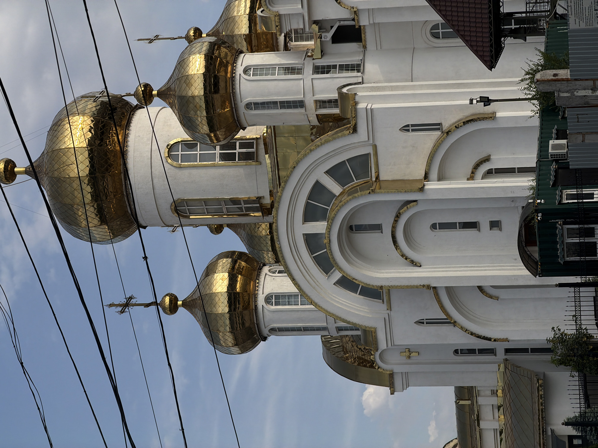 This is a photograph of a white building with golden domes, possibly a church. The image is oriented vertically, with the building's facade stretching upwards. Several golden domes are visible, some with ornate cross finials. The building features arched windows and decorative gold trim along its exterior. Power lines are strung across the sky in the foreground, obscuring parts of the building and sky. The sky appears to be partly cloudy, suggesting daytime. The architecture suggests a religious building, likely in a style common in Eastern Europe or Russia. No people are visible in the image. There is no discernible text within the frame.