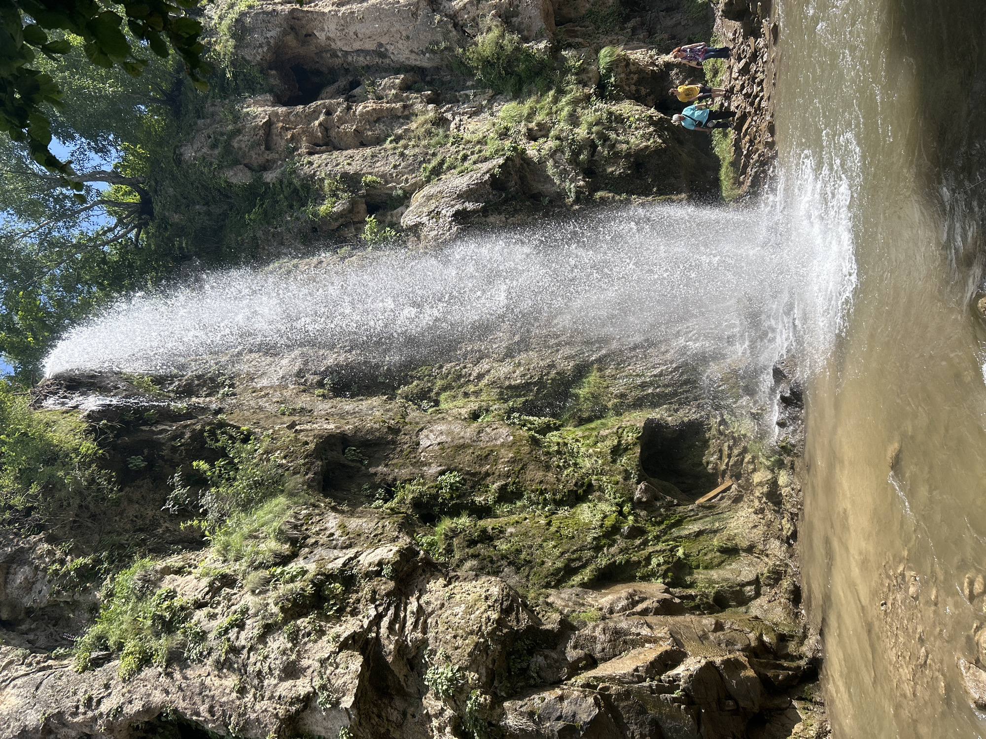 In the city of Krasny Kurgan, Russia, a powerful waterfall cascades down a sheer rock face. The water, appearing as a white sheet, plunges into a murky pool at the base. The rocky cliffs are adorned with lush green vegetation, clinging to the crevices and overhangs. Sunlight dapples the scene, illuminating the spray from the waterfall. On the rocky bank to the right of the waterfall, three individuals are visible. A person in a blue shirt and black pants stands closest to the waterfall, facing towards it. To their left, a person in a yellow t-shirt and dark shorts is also facing the waterfall. Further to the right, a person in a purple and white patterned shirt and blue jeans stands with their back partially turned, observing the scene. The environment is natural and rugged, with rocks and sparse vegetation lining the water's edge.