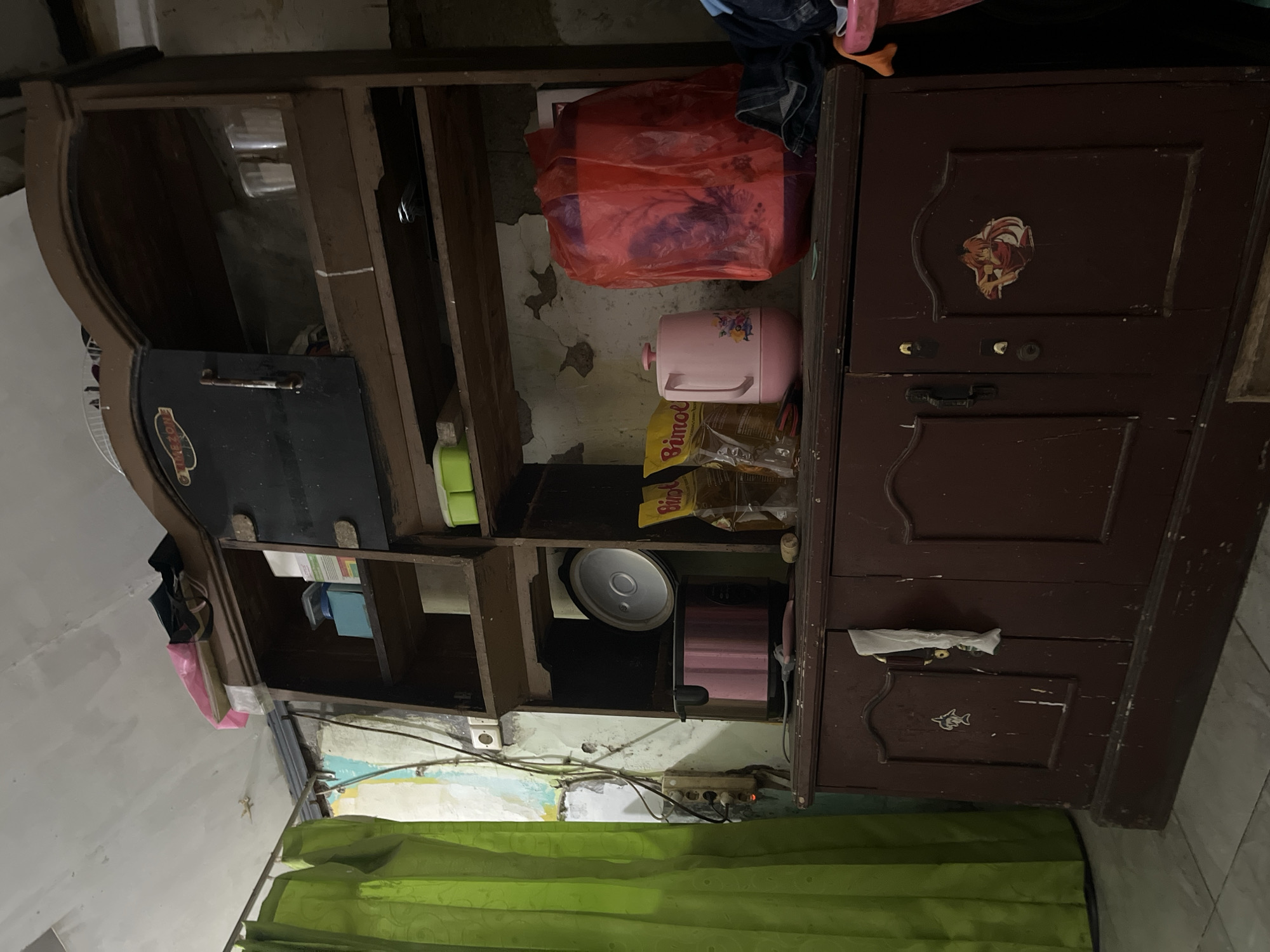 The image depicts a cluttered room, possibly a kitchen or storage area, in Pekalongan, Indonesia. A large, dark wooden cabinet with glass doors dominates the right side of the frame. Inside the cabinet are various items, including a pink rice cooker, two bags of cooking oil, a pink kettle, and a large red plastic bag. The cabinet doors are closed, revealing a sticker of an anime character on the right door and a white cloth hanging from the left door. The walls are weathered, with peeling paint and visible electrical wiring. A green curtain hangs on the left side of the room, partially obscuring a window. The floor appears to be tiled. The overall impression is one of a lived-in space, with items placed casually and without particular order.