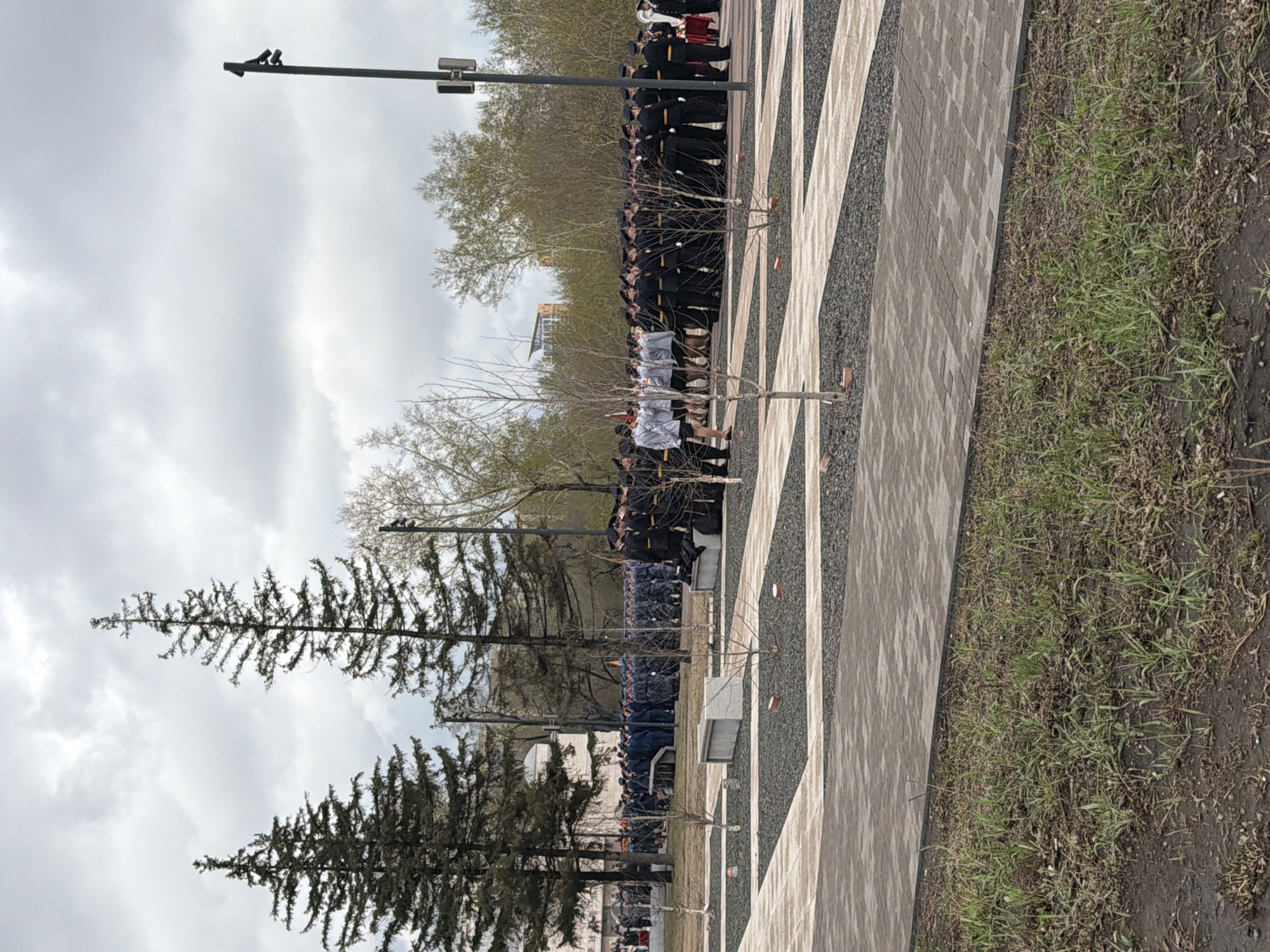 The image shows a scene in Krasnoyarsk, Russia. A line of people, predominantly dressed in dark uniforms, stands in a row along a walkway with a patterned surface. A smaller group of people in white attire is interspersed within the line. In the background, trees without leaves are visible, and a city building is partially seen. To the left are tall evergreen trees. A streetlight with multiple fixtures is present. The ground adjacent to the walkway is covered with grass and bare earth. The sky is overcast with grey clouds.