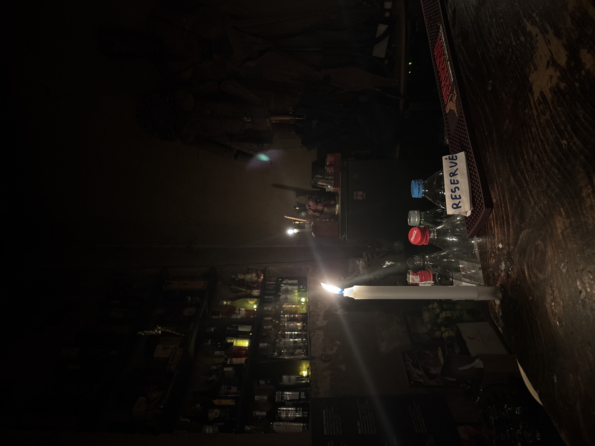 The image depicts a dimly lit interior scene, likely a bar or pub in Sofia, Bulgaria, with a primary light source being a single burning candle. On the right side of the frame, a dark wooden bar counter with a visible grain serves as the main surface. A tall, white candle stands upright on this counter, its bright flame casting significant light and flares into the dark surroundings.
Adjacent to the candle, several plastic bottles are placed on the counter; one is clear with a blue cap, and another has a red Coca-Cola cap. A small, white rectangular card is propped up against these bottles, bearing the word "RESERVE" handwritten vertically in blue ink. Further along the counter, a red and black textured bar mat is visible, clearly displaying part of the "JOHNNIE WALKER" brand name.
In the background and on the left side of the image, dimly lit shelves are stocked with numerous glass bottles of various shapes and sizes, suggesting an array of alcoholic beverages. The low light conditions indicate it is likely evening or night, creating a rather intimate and cozy atmosphere, potentially due to a power outage or a conscious choice for ambient lighting. No people are visible within the frame.