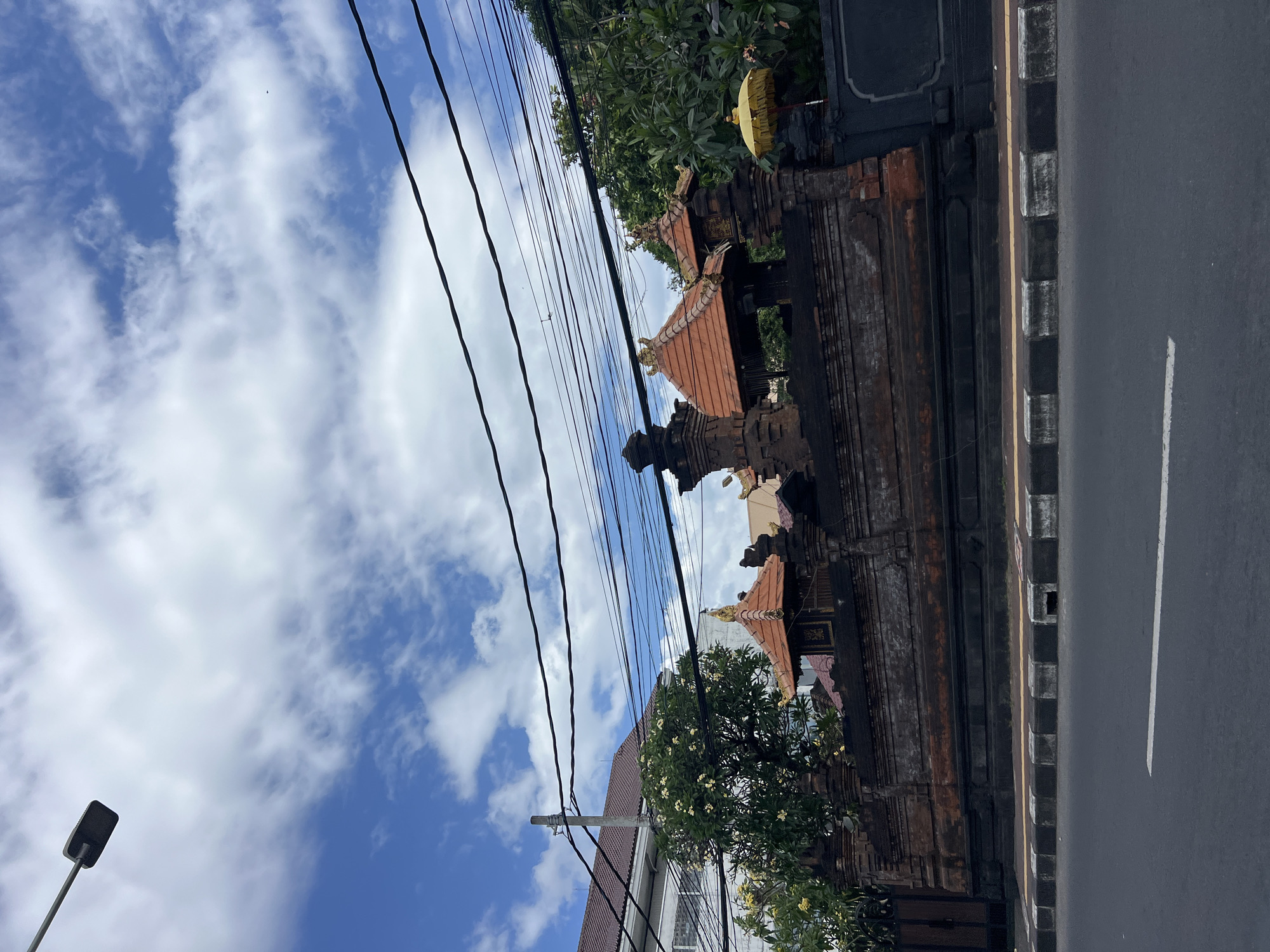 The image depicts a street scene in Denpasar, Indonesia, under a partially cloudy blue sky. A traditional Balinese-style wall, constructed from dark brick with ornate stone carvings, stretches across the frame. Several tiered roof structures with red tiles and gold accents emerge from behind the wall, characteristic of Balinese temple architecture. A large tree with small yellow flowers is visible behind the wall on the left side of the frame. Overhead, a dense network of black electrical wires is strung between poles. A street lamp extends from the upper left corner of the image. The foreground shows a paved road with a white line marking a lane. The curb is a combination of black and white painted concrete blocks. No people are visible in the image. The overall impression is one of a cultural and urban intersection with clear Indonesian architectural influences.