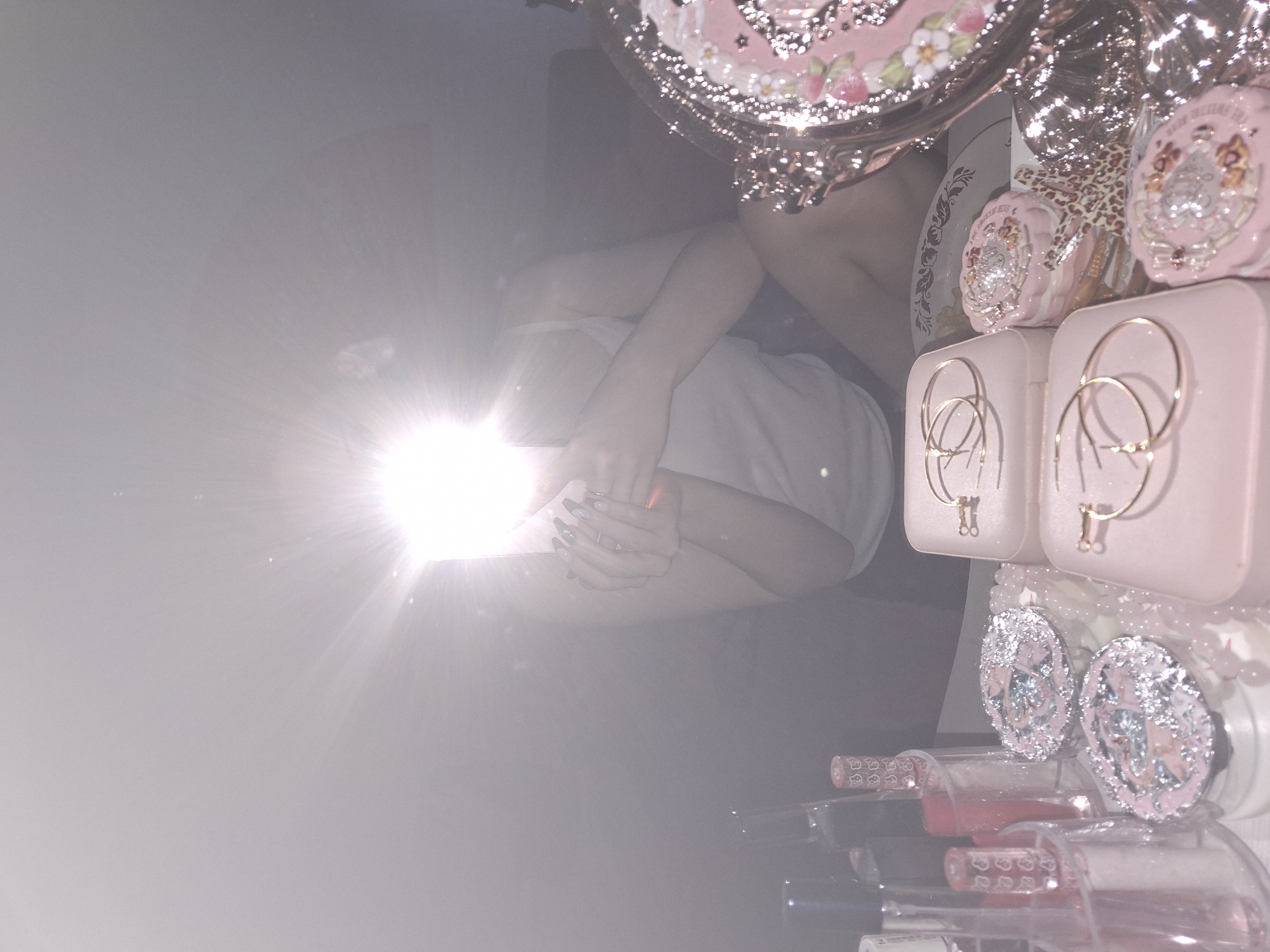 A bright flash obscures most of the scene. A person wearing a white top and light skin tone is partially visible, seated and hugging their knees. Above them, there are several pink and silver items with a decorative, possibly "girly," aesthetic. These objects include jewelry, possibly earrings, and decorative containers. Below the person, there is an array of lipsticks or similar cosmetic tubes. The location in the city of Chaikovsky, Russia, is not discernible from this image.
