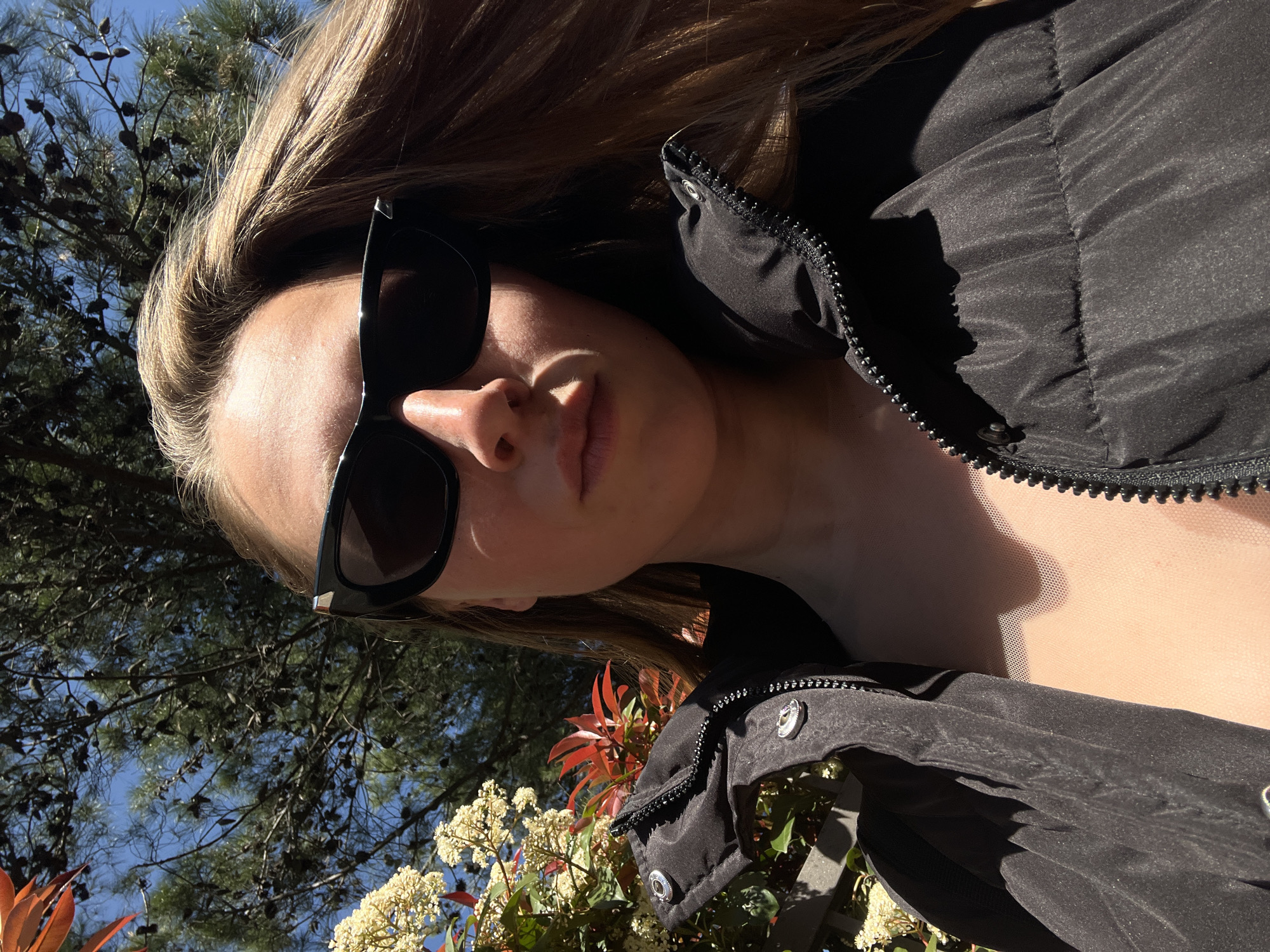 A young woman wearing sunglasses and a black puffer jacket poses outdoors. The background includes a tree and a blue sky. The presence of distinct plants suggests a garden or park setting. Due to the lack of landmarks, it is impossible to determine if the scene is specifically in Budva, Montenegro, without further visual clues or metadata.