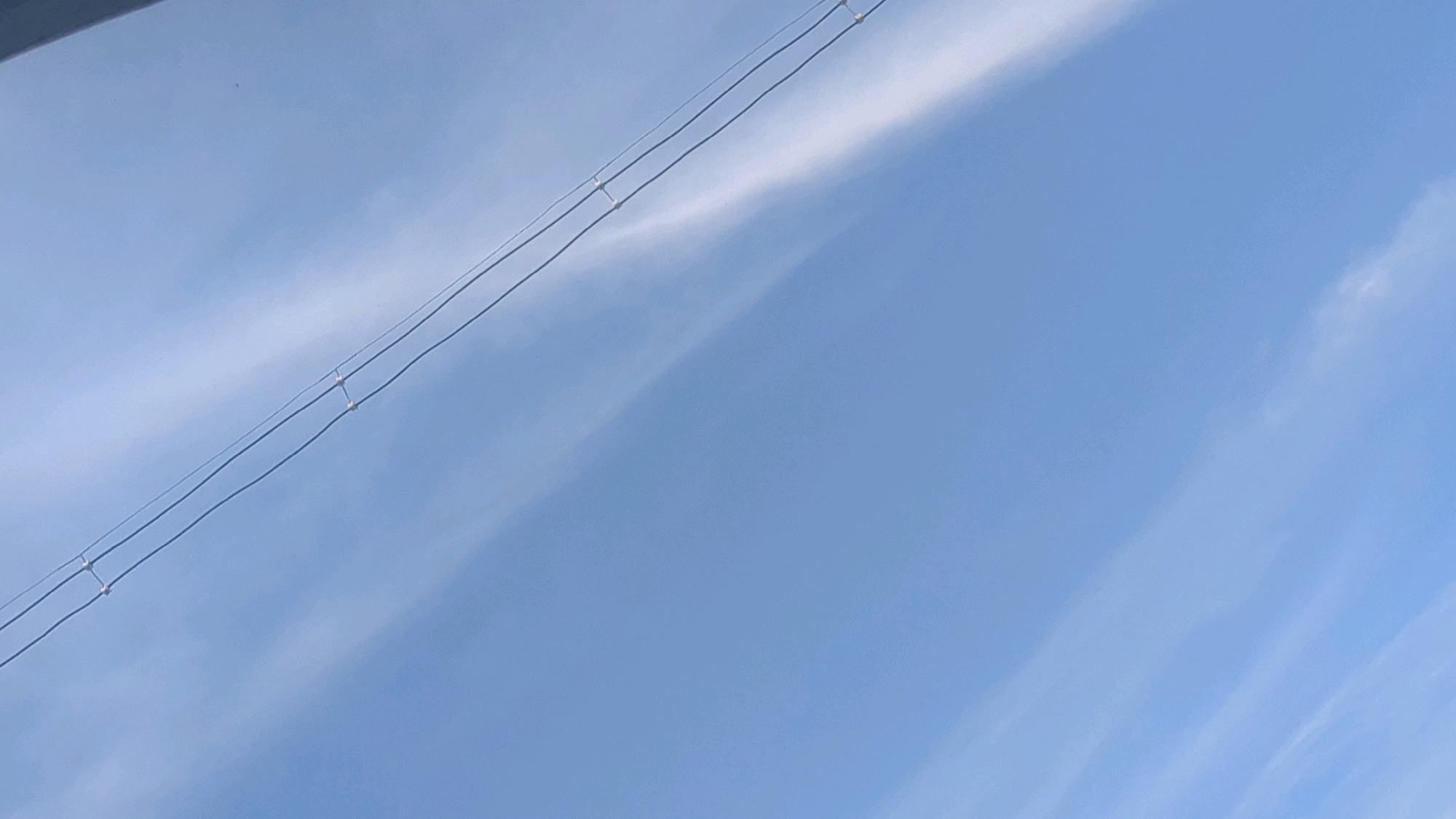 The image shows a clear blue sky with wispy clouds and power lines stretching diagonally across the frame. The power lines are thin and parallel, with small insulators connecting them. There is a hint of a dark structure in the upper left corner, possibly a rooftop or the edge of a building, suggesting an urban or suburban setting. The lighting indicates it is daytime, with the sun likely positioned above and to the right of the frame. There are no people or discernible activities visible in the image. The overall scene is tranquil and depicts a common urban sky view. The image is presumed to be from Alipur, India, based on the provided context.