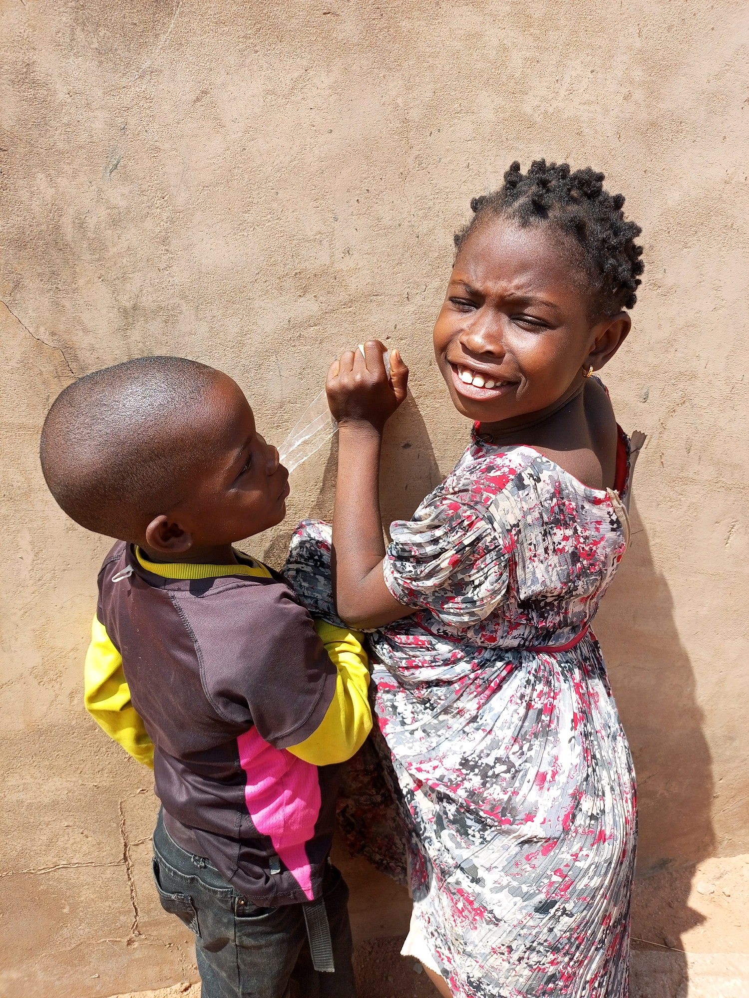 In Tempure, Nigeria, two children are captured against a textured, beige wall. A young boy, facing away from the camera and to the left, wears a dark brown and yellow jacket with a pink stripe down the back, paired with dark denim jeans. He appears to be interacting with something unseen on the wall. To his right, a young girl with dark, braided hair and a bright, patterned dress with red and white accents is facing the camera with a wide, toothy grin and her eyes squinted. The lighting suggests it is daytime, with shadows cast on the wall.