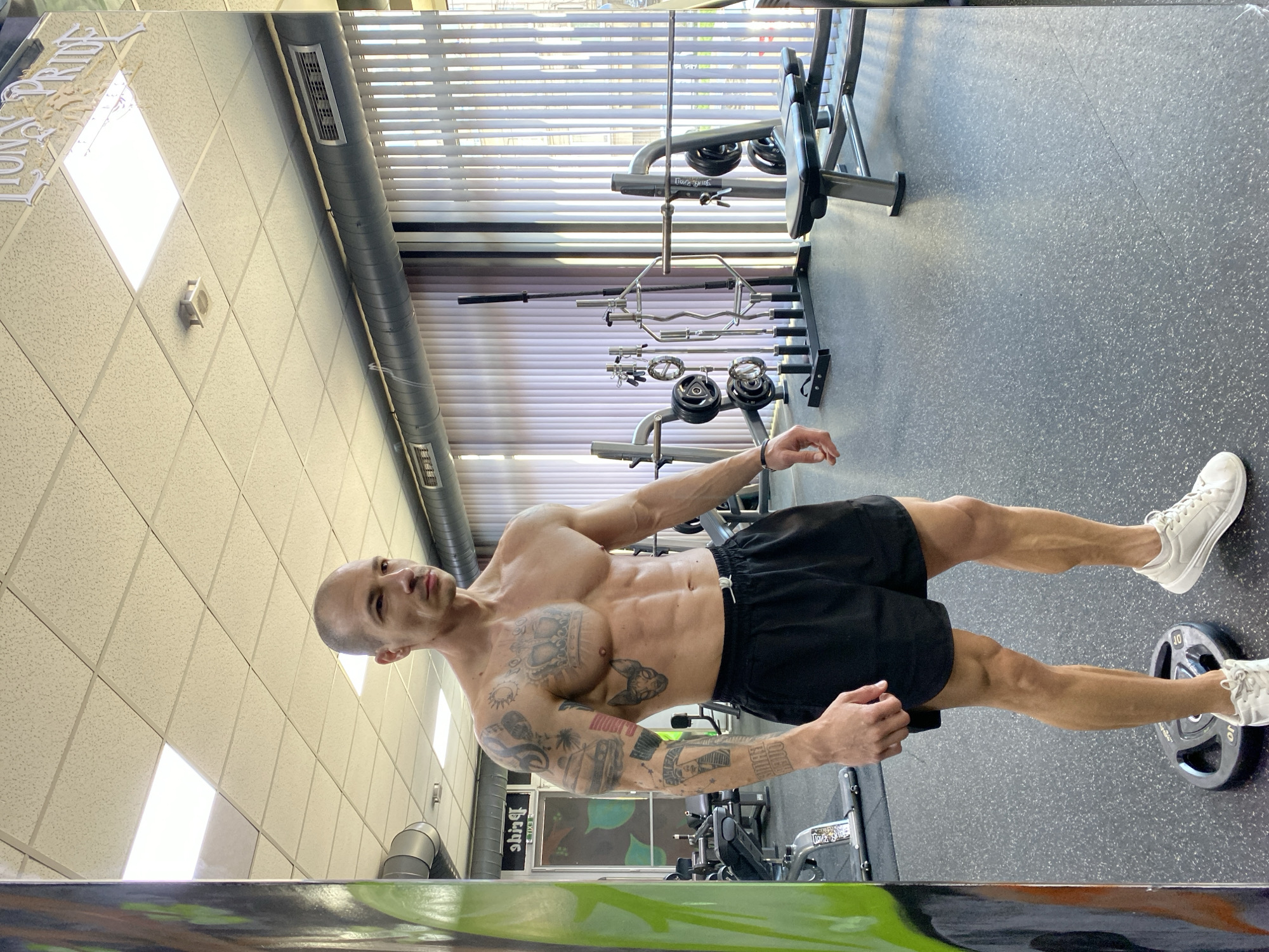 A medium-shot photo shows a muscular, shirtless man wearing black shorts and white sneakers in a gym. He is standing with one foot on a 10-pound weight plate, his right arm extended to the side. Tattoos are visible on his arms and chest. In the background are various pieces of gym equipment, including barbells and weight racks. The gym has a tiled ceiling with fluorescent lighting and windows with blinds. The location appears to be in the city of Pleven, Bulgaria, based on signage in the background.