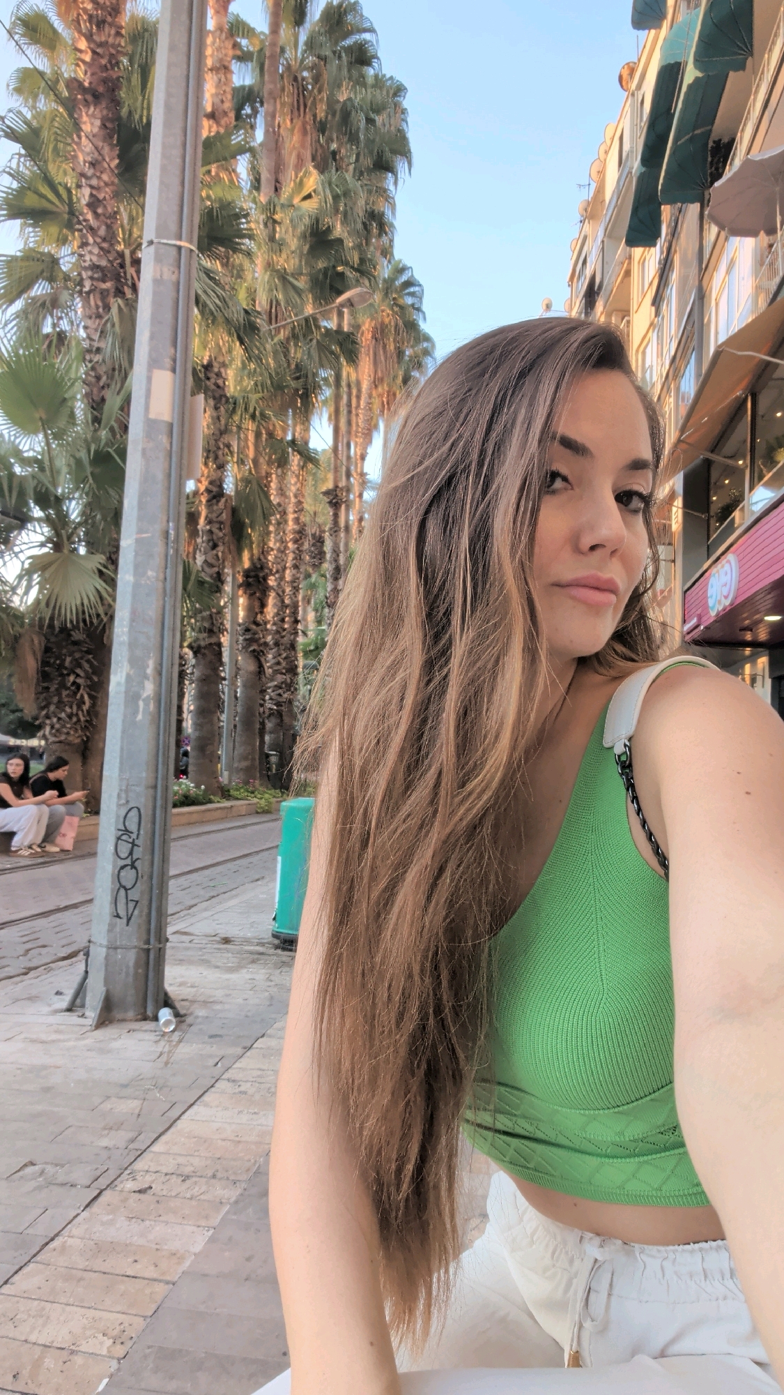 The image captures a young woman taking a selfie outdoors in Antalya, Turkey. She is wearing a vibrant green crop top and white pants. Her long, brown hair is casually styled and falls over her shoulder, partially obscuring her face. She is positioned in the foreground, with the background showing a street scene.
To her left, a prominent lamppost stands tall, bearing some graffiti. Beyond the lamppost, a row of tall palm trees lines the street, their fronds creating a tropical ambiance. A tram track is visible on the ground, indicating this is likely a public transportation route. Further in the background, a green trash bin is present, and two young women are seated on a bench, appearing to be engaged in conversation. The buildings on the right side of the image suggest an urban environment, with shops and awnings characteristic of a commercial street. The lighting indicates it is daytime, possibly late afternoon, with the sun casting warm tones on the scene.