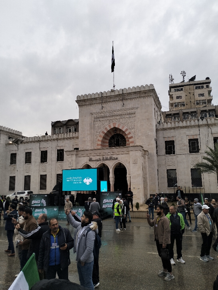 An outdoor scene depicts a crowd gathered in front of a large, light-colored building with Arabic script above the entrance. A black and white flag flies atop the building. A stage with screens displays an emblem resembling an eagle. People are milling around, some taking photos, and wearing casual attire. The weather appears overcast, and the ground may be wet.