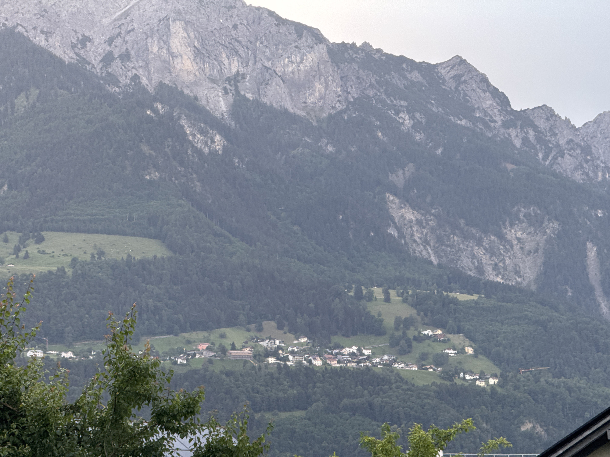 The image shows a panoramic view of the village of Nendeln, Liechtenstein, nestled in a valley at the base of a steep, rocky mountain range. The mountain slopes are covered in dense green forests, with patches of lighter green meadows interspersed. The village itself consists of numerous houses with red and white roofs, clustered together on the hillside. Some of the houses appear to be larger buildings, possibly hotels or community centers, while others are smaller residential homes. In the foreground, the tops of trees with green leaves are visible, partially obscuring the view of the village. The sky is overcast, giving the scene a somewhat muted and atmospheric feel. There are no people or active vehicles visible in the image.