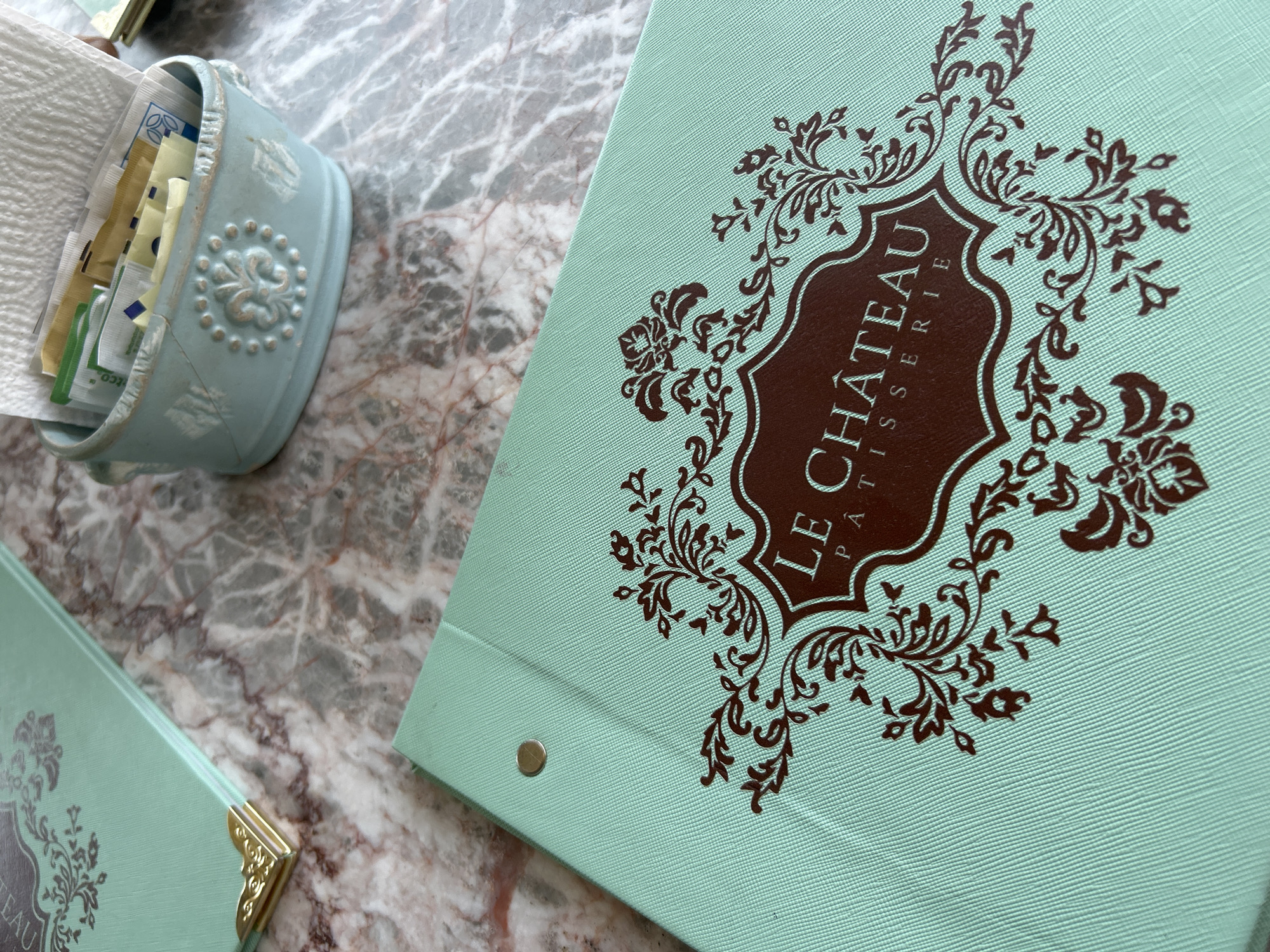 An overhead shot captures a light blue menu or binder resting on a marble-patterned tabletop. The menu is a prominent subject, featuring ornate dark brown floral and scrollwork designs around a central crest. Within this crest, the text "LE CHÂTEAU PÂTISSERIE" is vertically aligned, with "LE CHÂTEAU" in a larger, elegant font and "PÂTISSERIE" in smaller, stacked letters below it. The menu cover has a textured, possibly fabric-like surface, and a silver-colored stud is visible near its spine on the left side.
To the upper left of the menu, a light blue, distressed container with decorative embossed patterns holds an assortment of individual sugar and sweetener packets of various colors (yellow, green, white, blue). A napkin is also partially visible next to this container. In the bottom left corner of the frame, another similar light blue menu is partially visible, distinguished by a decorative gold metallic corner protector. The background consists entirely of the mottled grey and brown marble surface of the table. The scene suggests an indoor setting, likely a patisserie or café, with items typically found on a table when ordering or dining.